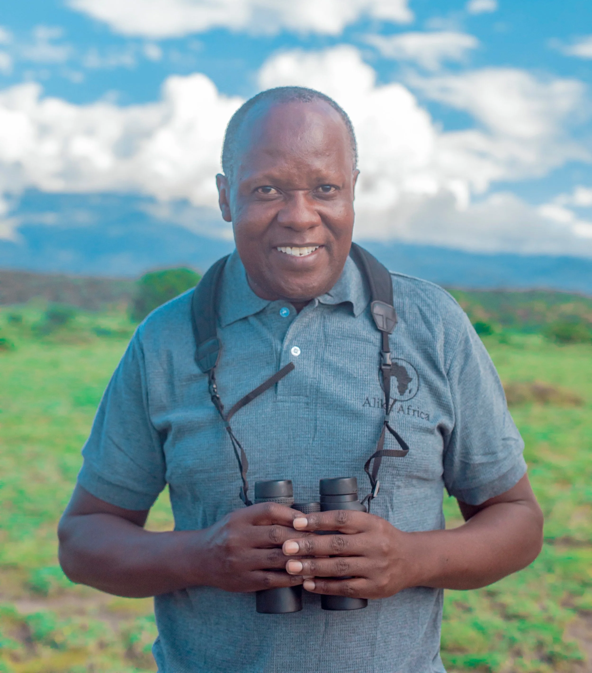 Person holding binoculars in a grassy field with a mountain and cloudy sky in the background.