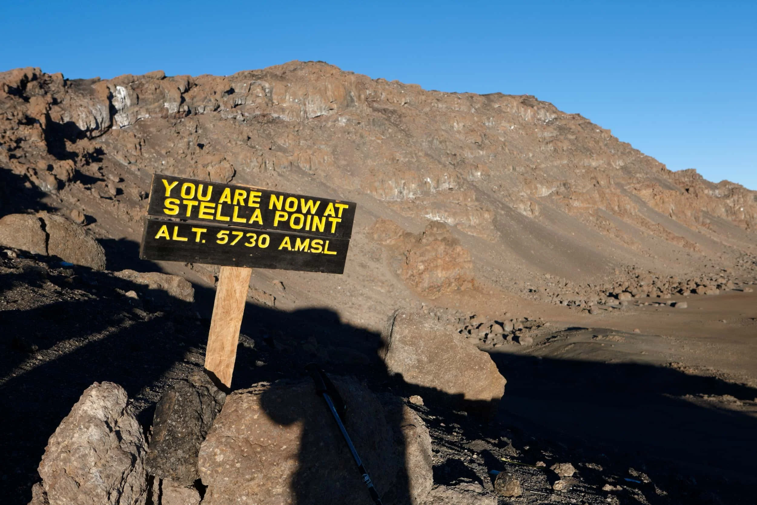 Sign indicating current location at Stella Point on Mount Kilimanjaro, elevation 5730 meters above sea level, in a rocky, mountainous landscape with clear blue sky.