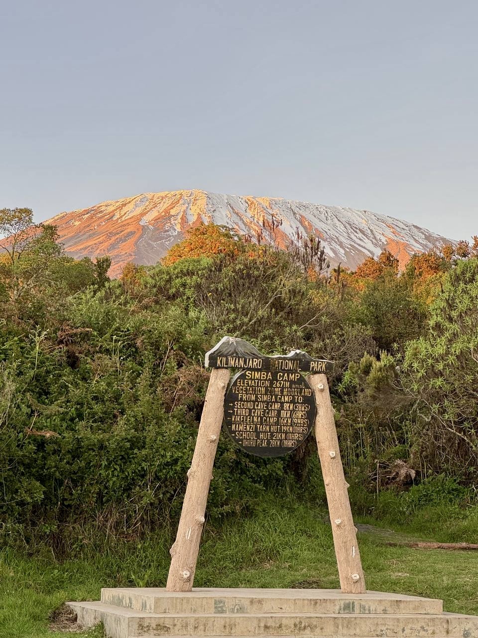 Volcano with colorful streaks on its slopes, forested area in the foreground, and a sign at Kilimanjaro National Park indicating hiking routes and elevation details.
