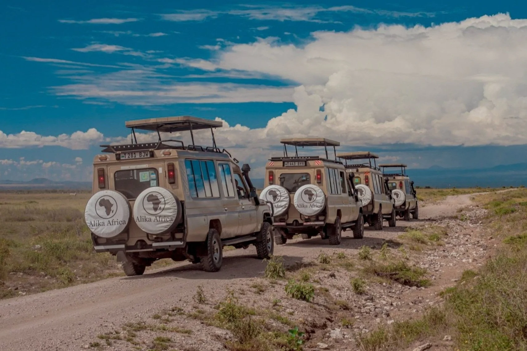 A row of safari vehicles driving on a dirt road in a savannah landscape under a partly cloudy sky, with "Alika Africa" branding on spare tire covers.