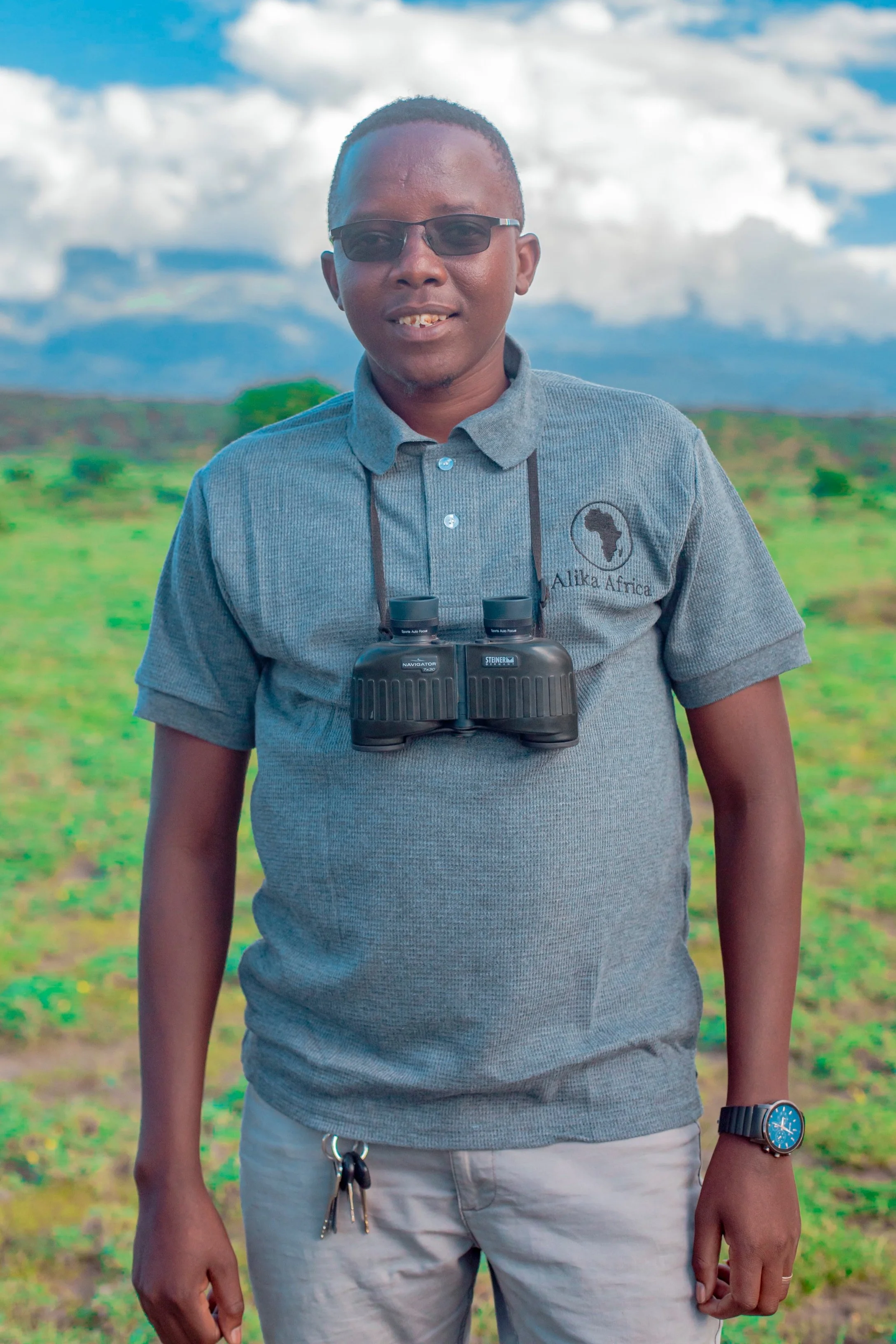Person in a gray polo shirt with binoculars around their neck, standing outdoors in a green field with a cloudy sky.