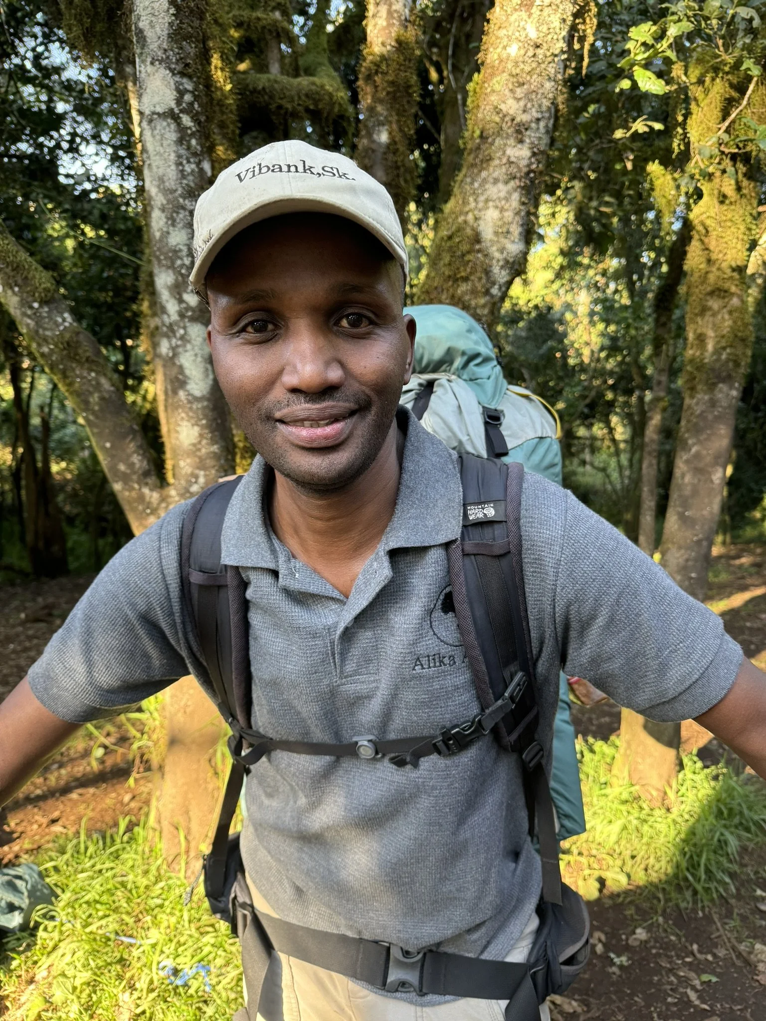 Man wearing a gray polo shirt and khaki cap, standing outdoors with trees in the background, carrying a backpack.