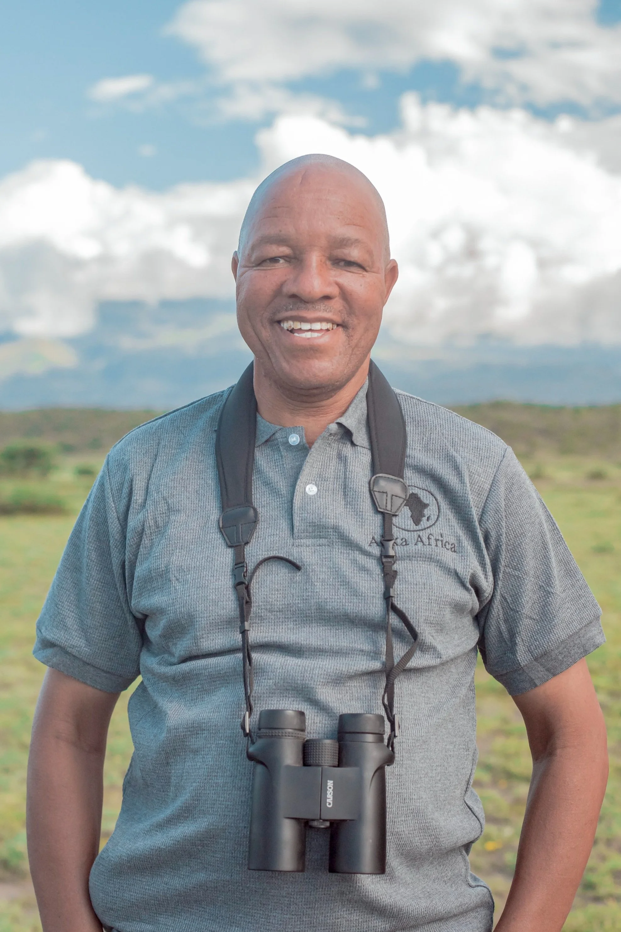 Man standing outdoors wearing a gray shirt and binoculars around his neck, smiling with a cloudy sky and grassy landscape in the background.