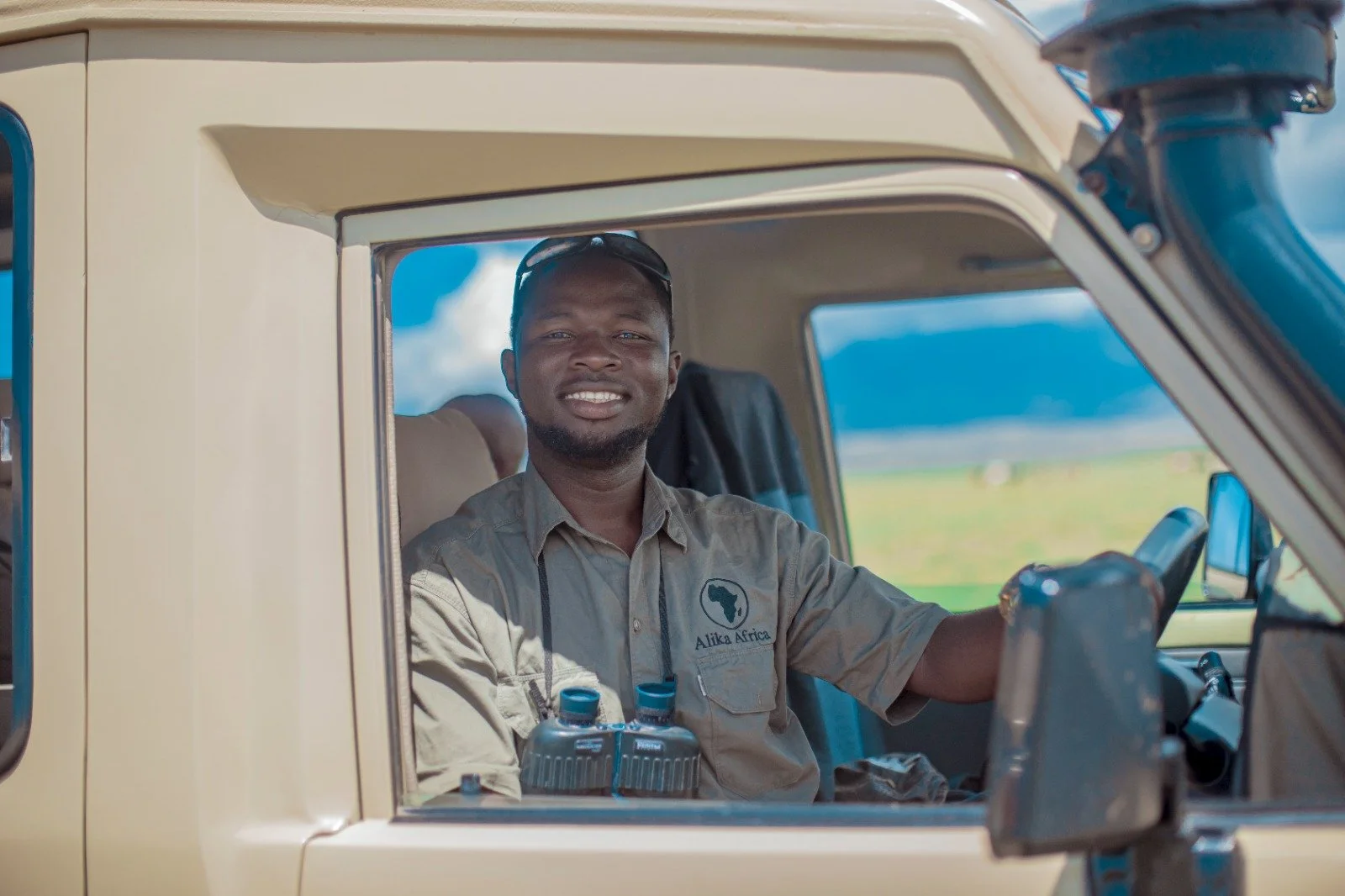 A smiling man sitting in the driver's seat of a safari vehicle, wearing a beige shirt with 'Alika Africa' logo, with binoculars on his chest, in a scenic outdoor setting with blue skies and green fields.