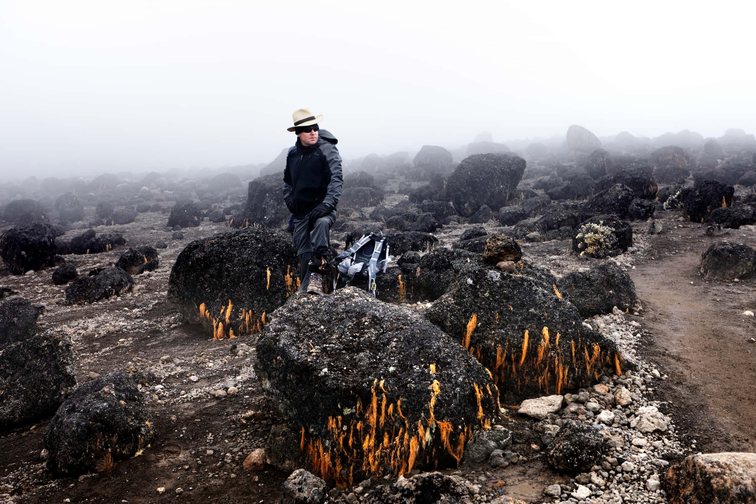 A person wearing a wide-brimmed hat, sunglasses, and dark outdoor clothing stands on a rocky, lava-covered terrain with orange lava tubes, surrounded by fog in the background.