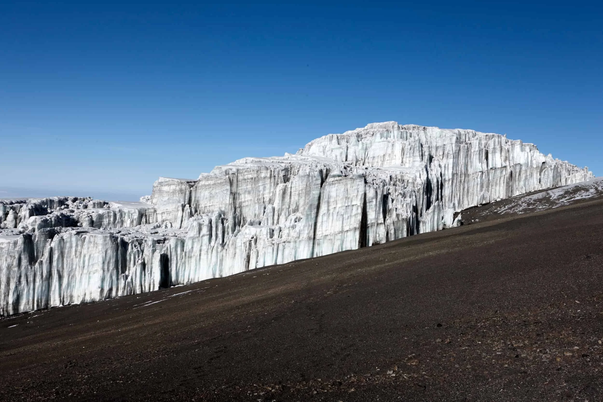 A large, white glacier with vertical ice formations and deep crevasses, under a clear blue sky, with dark, rocky ground in the foreground.