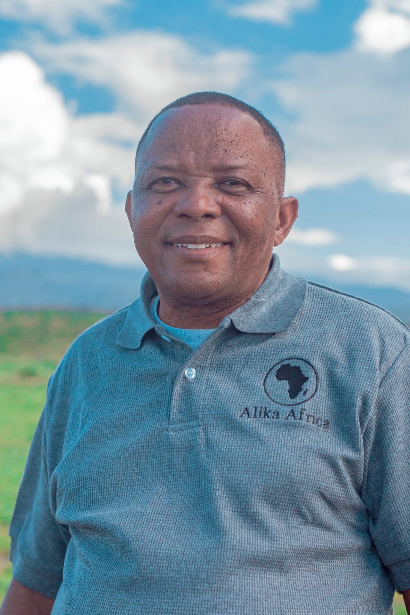 Man wearing grey shirt with Alika Africa logo, standing outdoors, blue sky and green field in background.