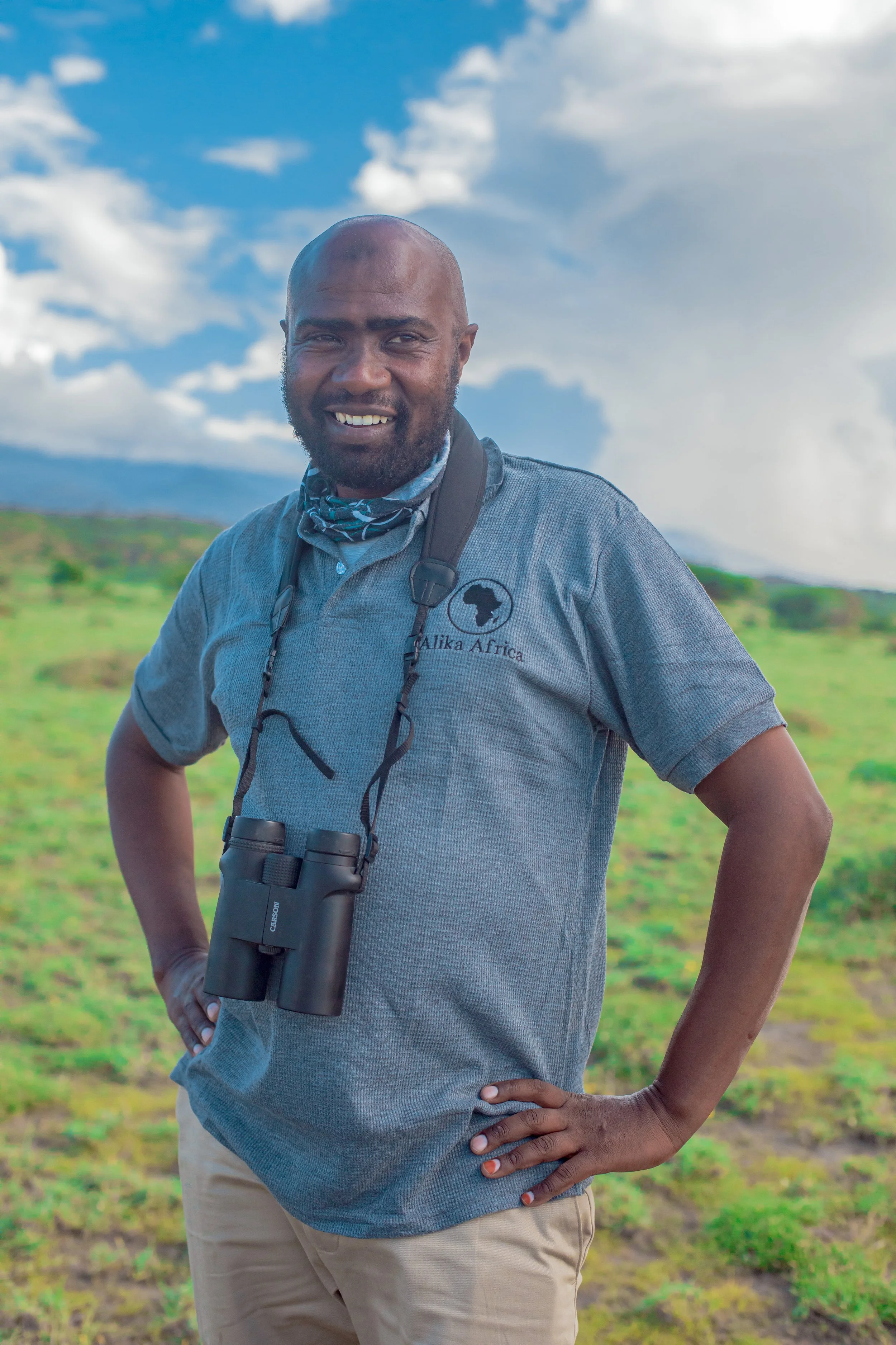 Smiling person outdoors with binoculars, in a green field under a blue sky with clouds.