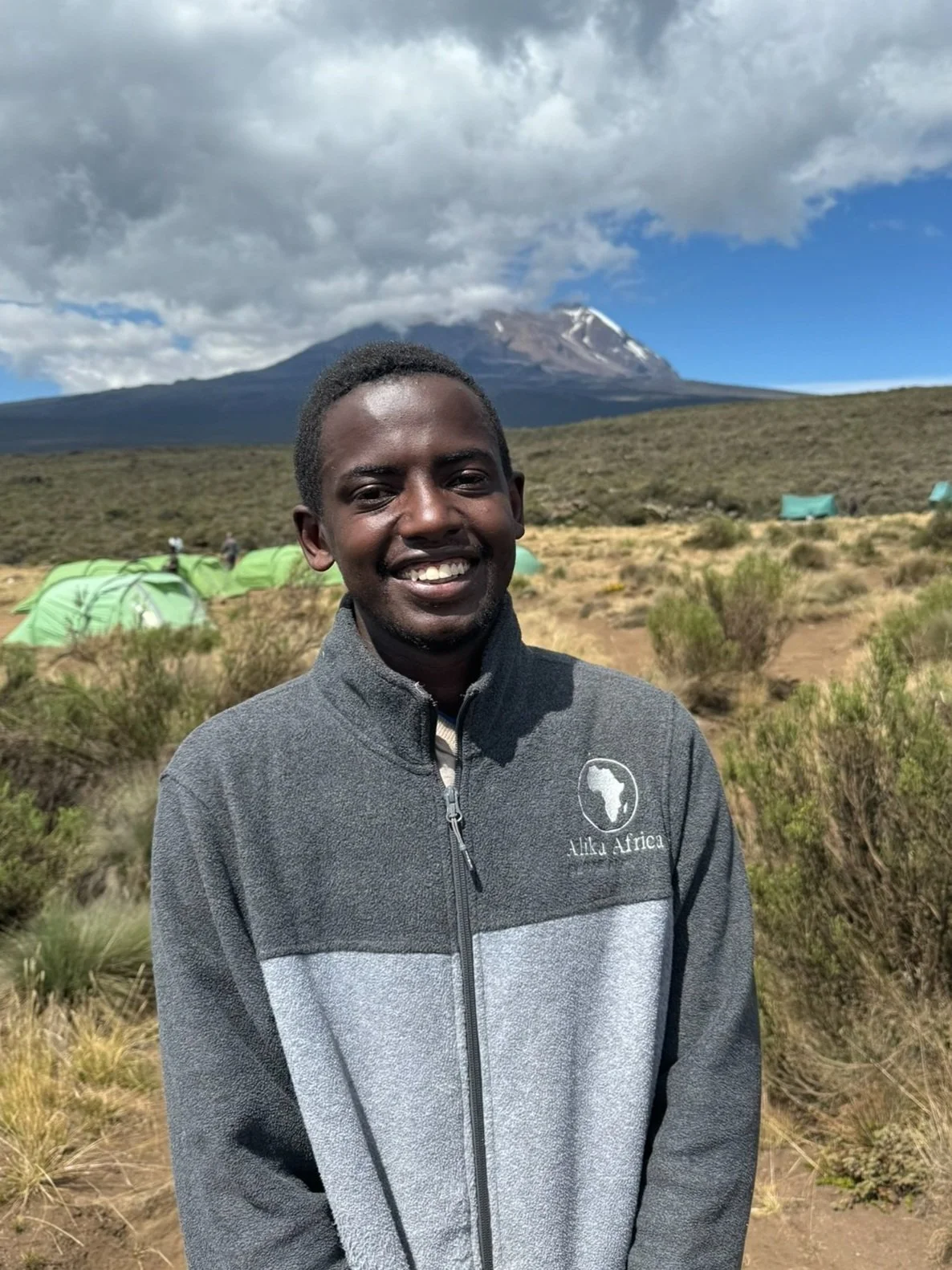Smiling person in outdoor setting with mountains and green tents in background.