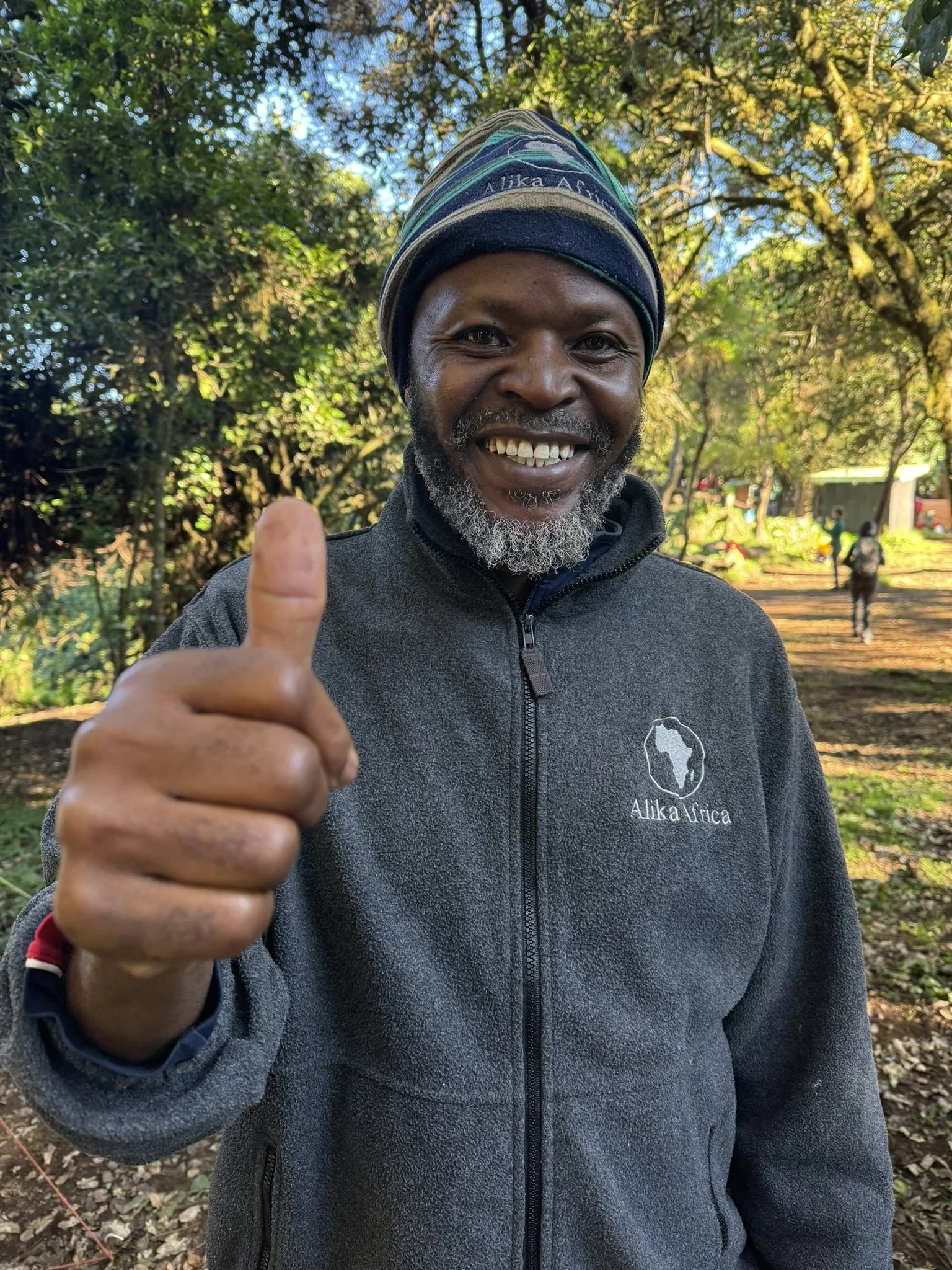 A person smiling and giving a thumbs-up outdoors, wearing a beanie and a gray jacket with "Alika Africa" on it, surrounded by trees.