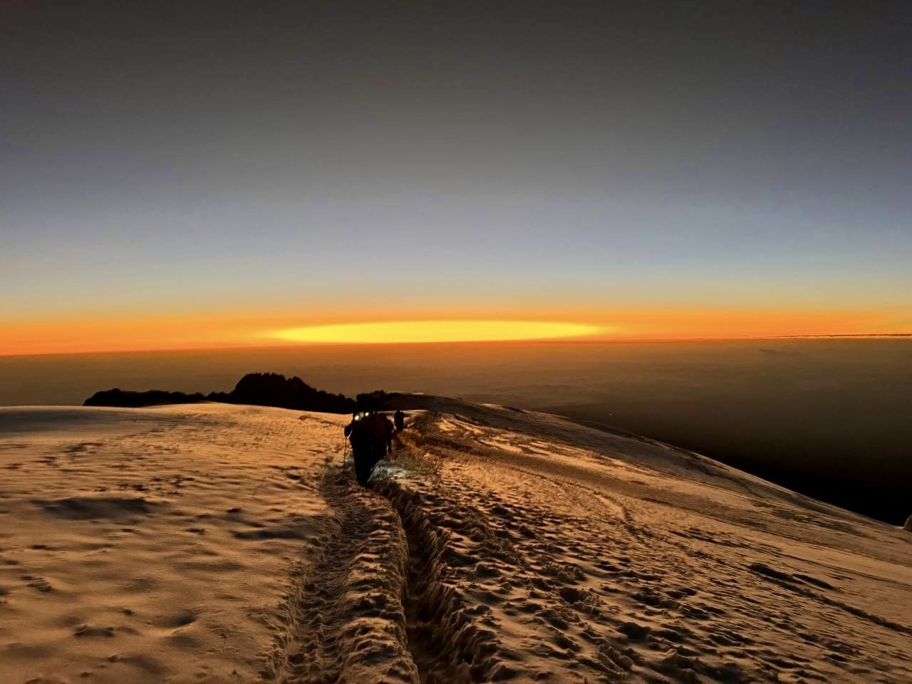 Hikers with headlamps walking along a snow-covered trail on a mountain at sunset, with the sky transitioning from orange to dark blue.