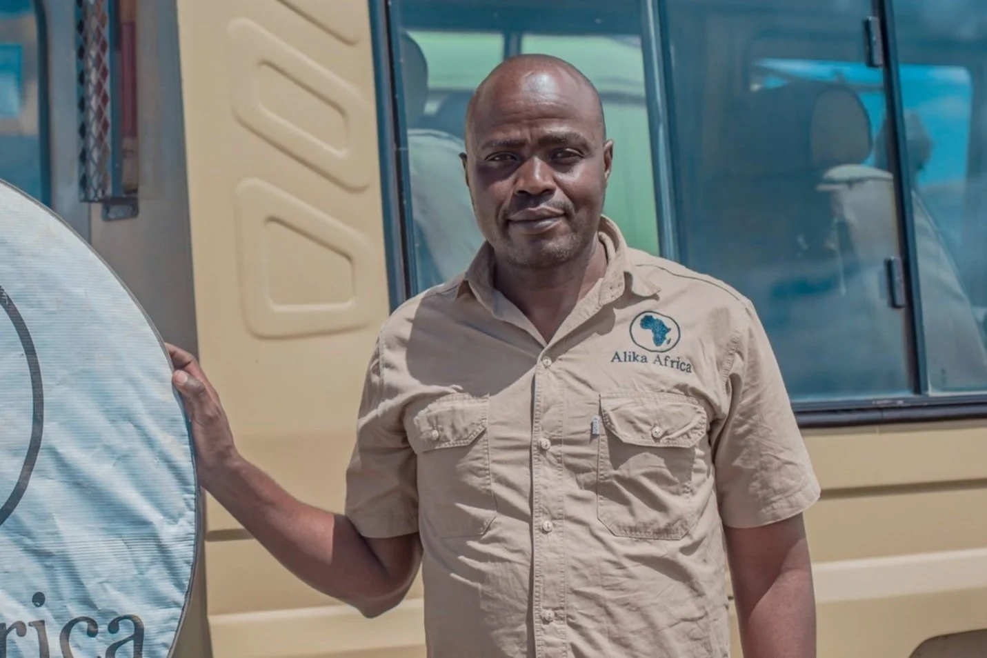 Man standing in front of a safari vehicle, wearing a beige shirt with 'Alika Africa' logo.