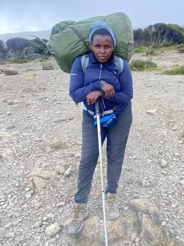Person hiking on a rocky trail, wearing a blue jacket, black pants, beanie, and hiking boots, carrying a large green backpack and holding a walking stick.