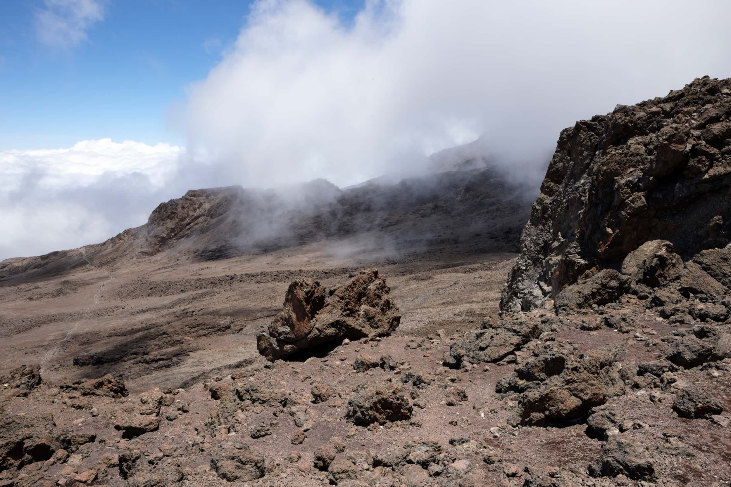 A barren, rocky volcanic landscape with clouds around the mountain peaks in the background.