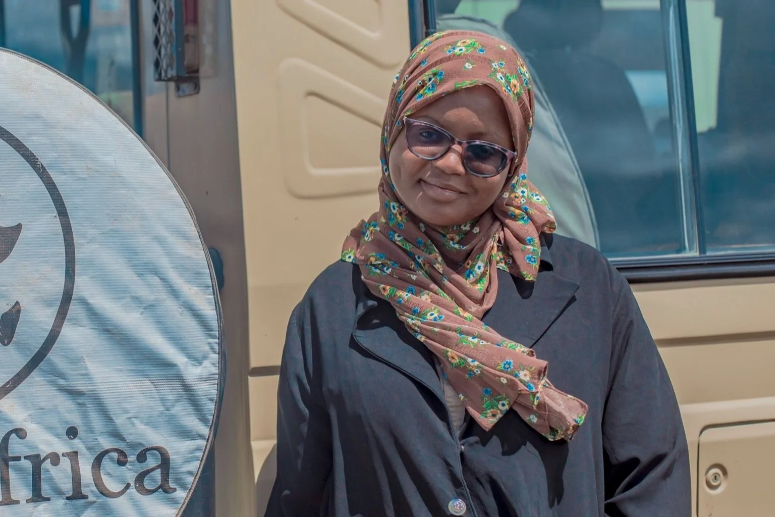 Person wearing a floral headscarf and sunglasses, standing next to a vehicle showing part of a tire cover and door, with the word "Africa" visible.