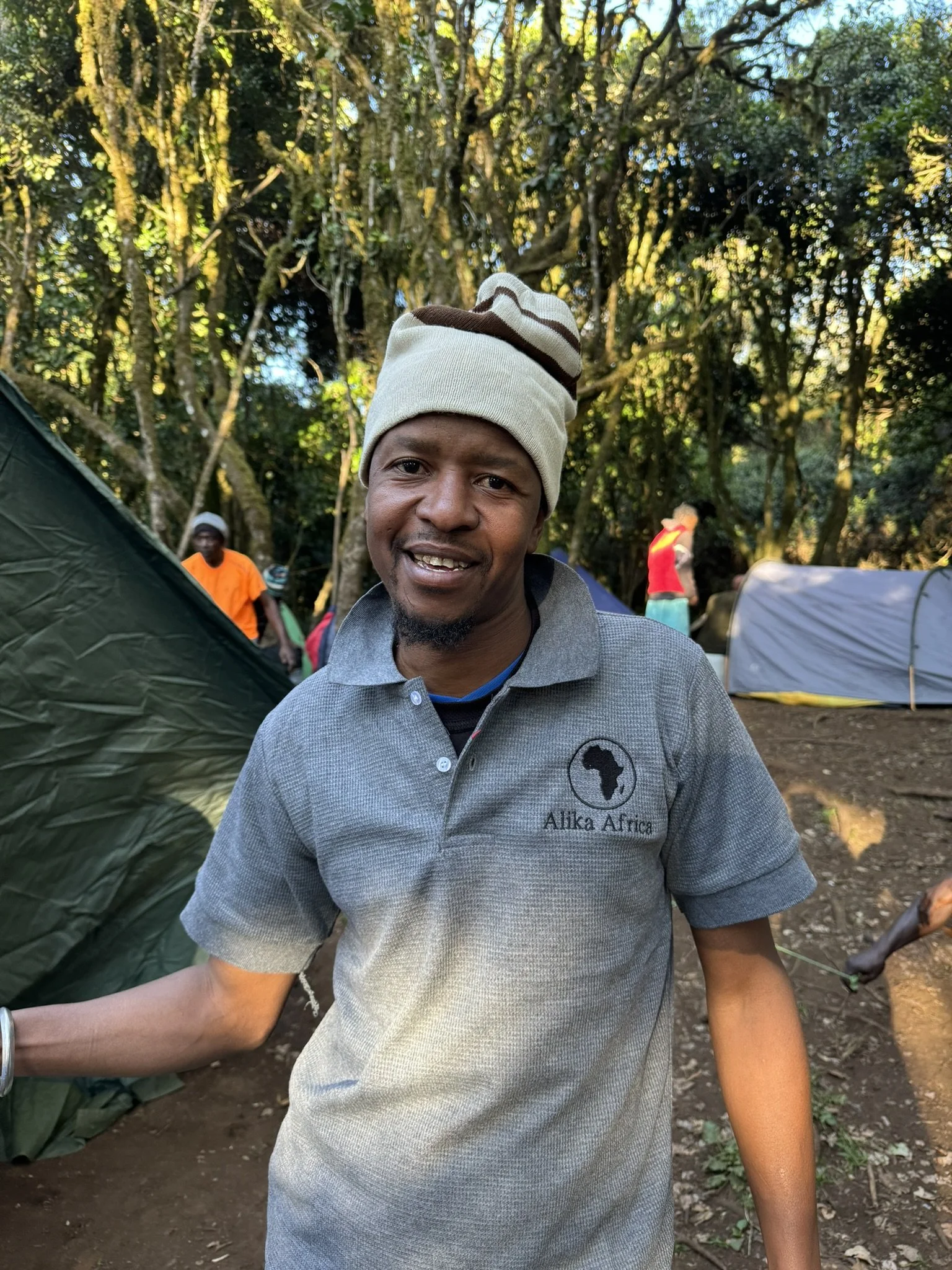 Man wearing a beanie and gray polo shirt standing outdoors in a wooded campsite with tents and trees in the background.