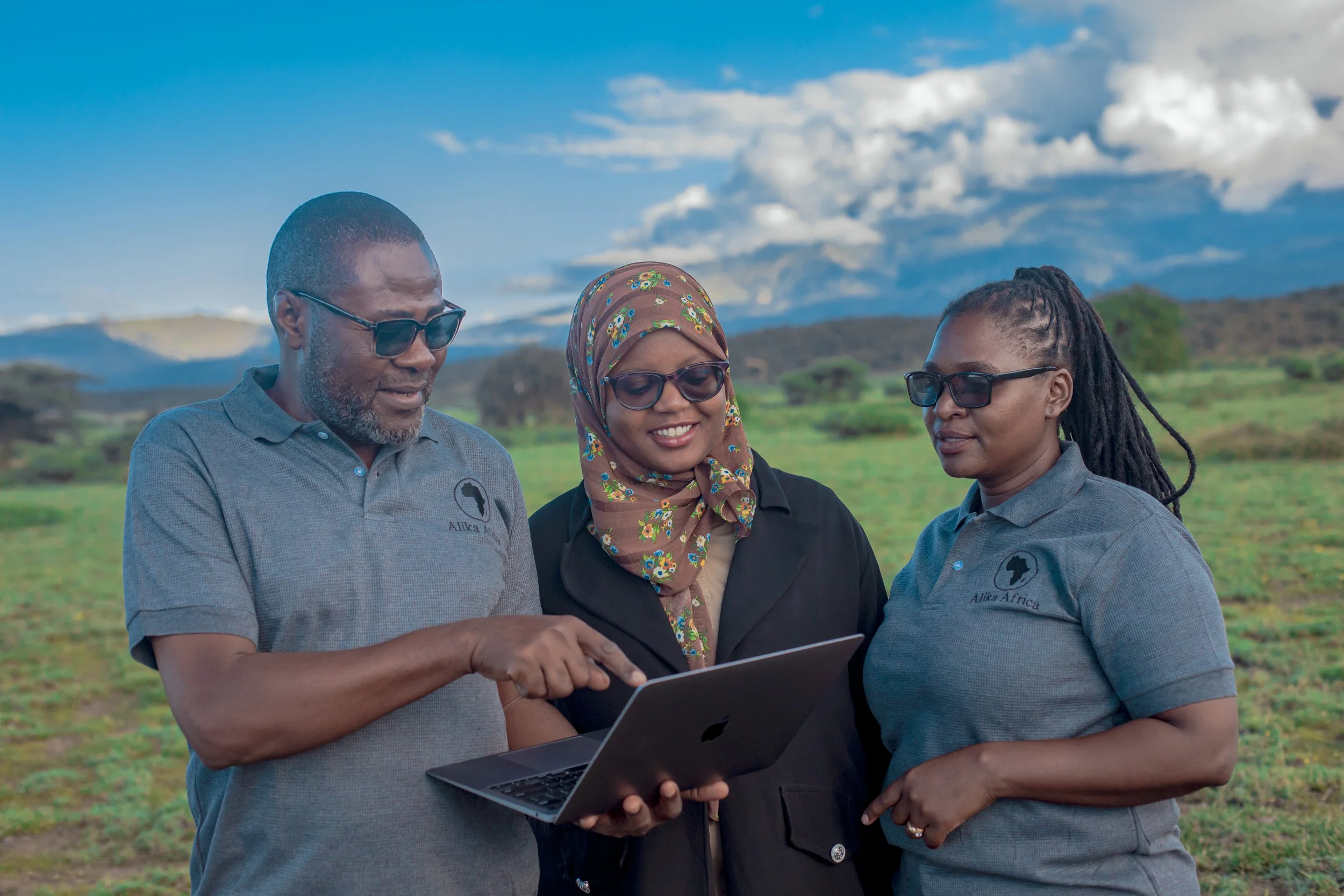 Three people standing in a grassy field looking at a laptop, with mountains and blue sky in the background.
