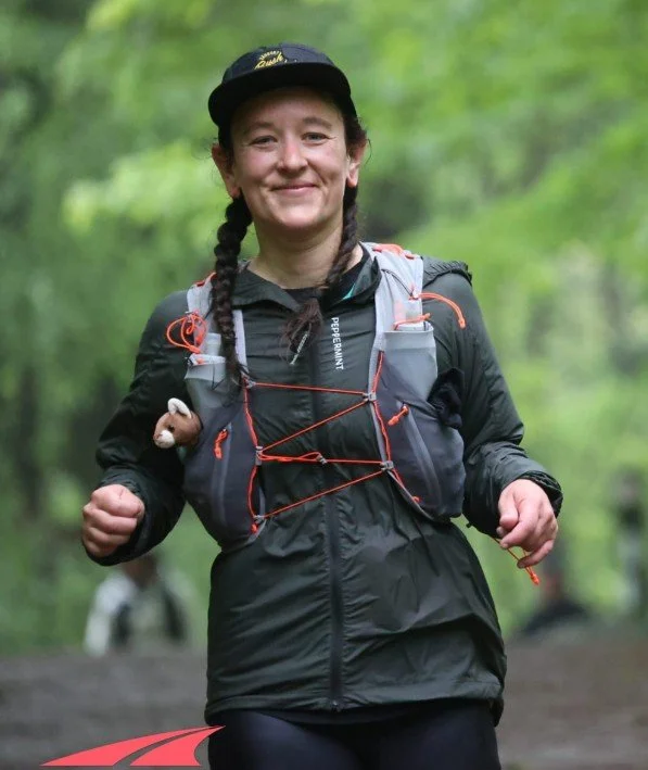 A woman jogging outdoors in a forest, wearing a black jacket, a cap, and a backpack with hydration tubes, smiling at the camera.