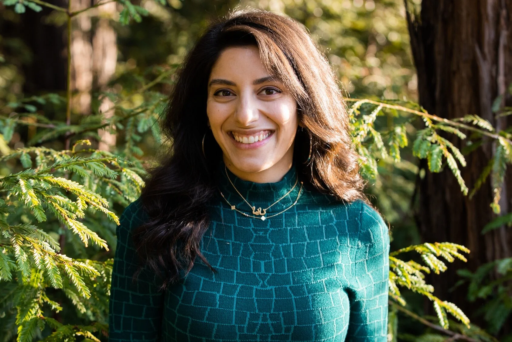Tania stands in a forest of redwood trees with a large smile and a green dress.