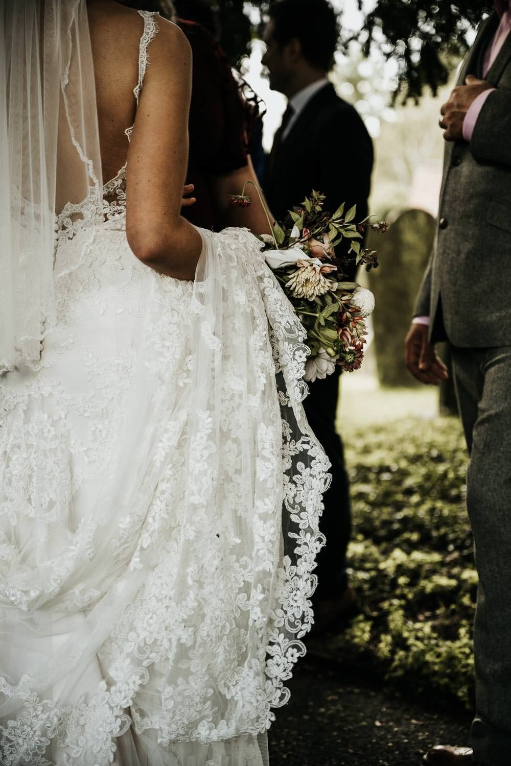 Lace wedding dress being held up by bride as she walks through church yard holding flowers