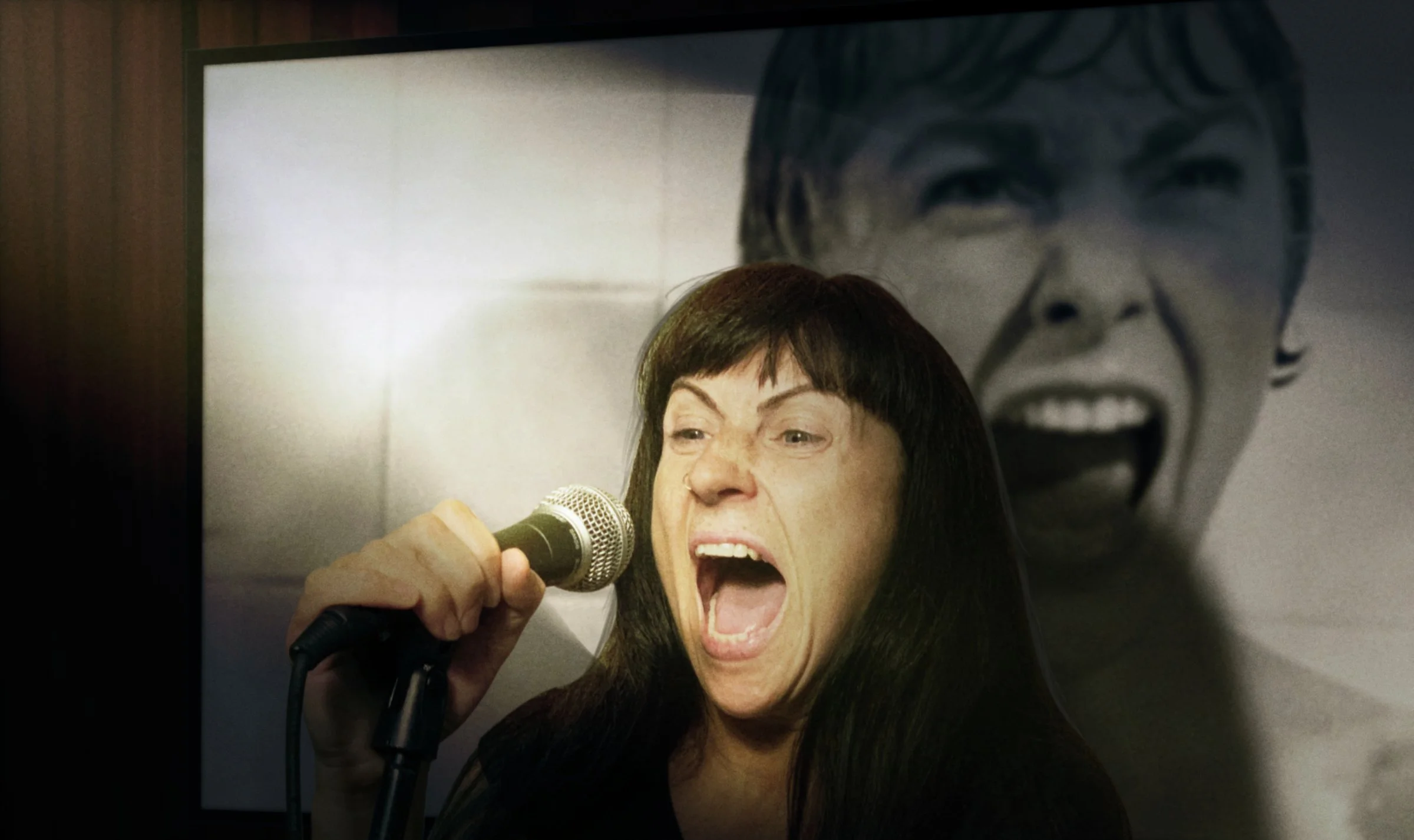 Close-up of a woman with long dark hair passionately singing or shouting into a microphone, with a large black and white poster of a face in the background.