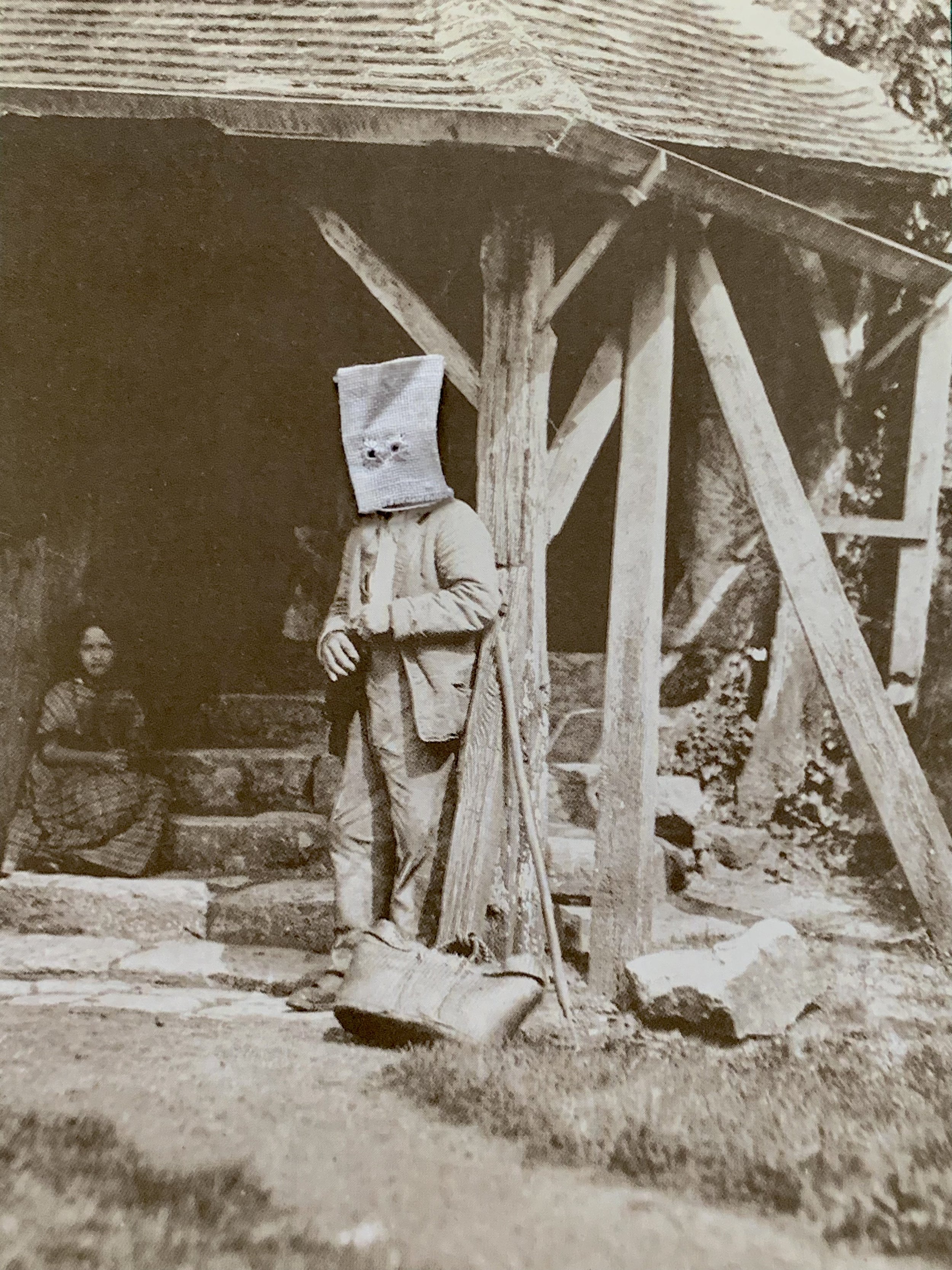 Vintage photo of a person standing outside a rustic house with a cloth sack over their head, and another person sitting on the steps next to them.