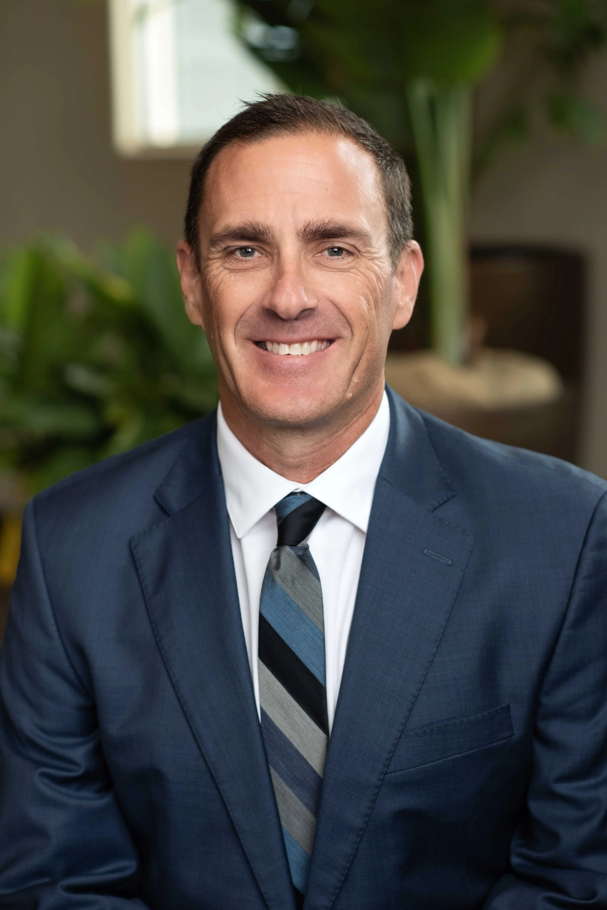 A man in a navy blue suit, white shirt, and striped tie smiling and looking at the camera, with a green plant and a window in the background.