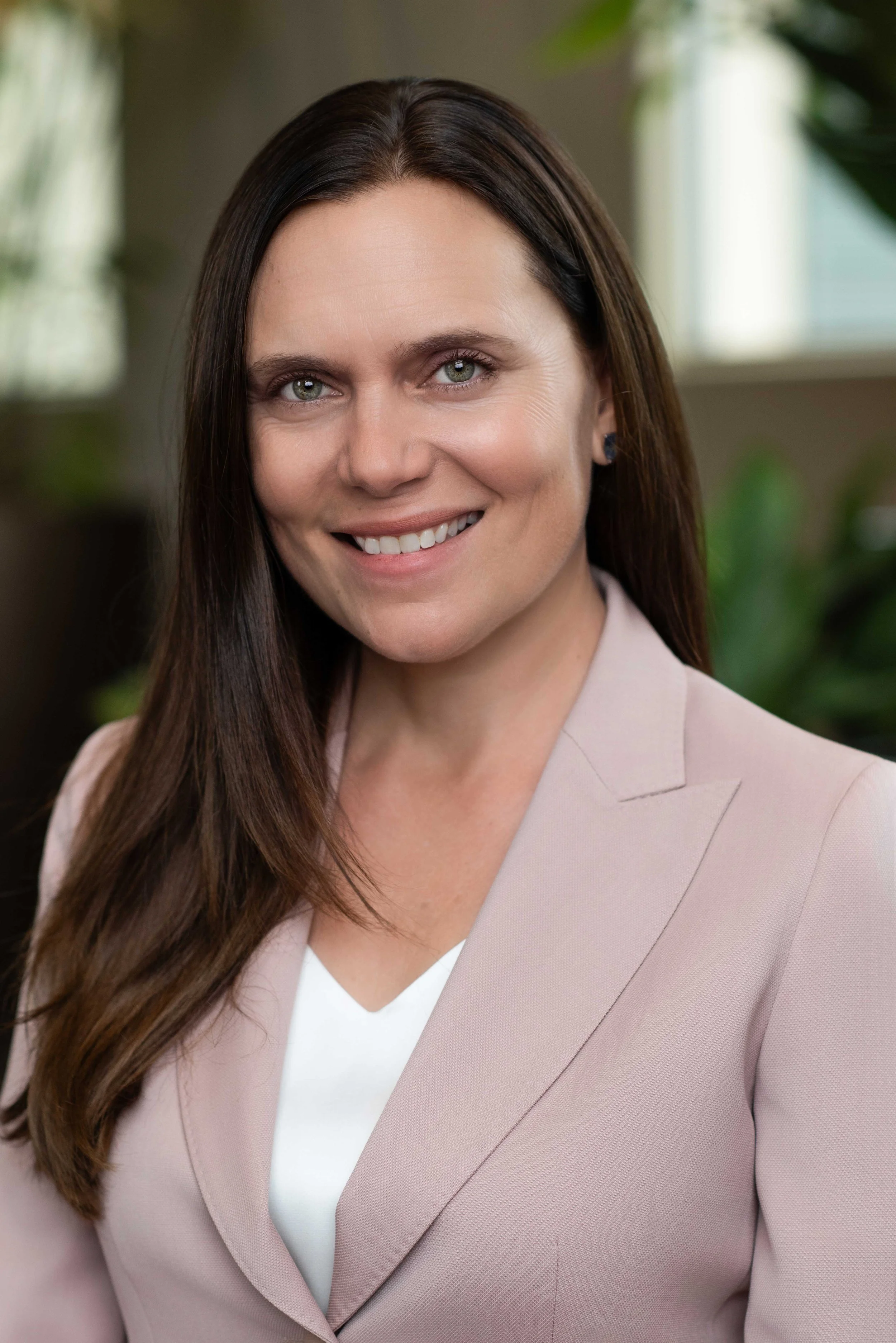 A woman with long brown hair and green eyes smiling, wearing a beige blazer and white top, in an indoor setting with greenery in the background.