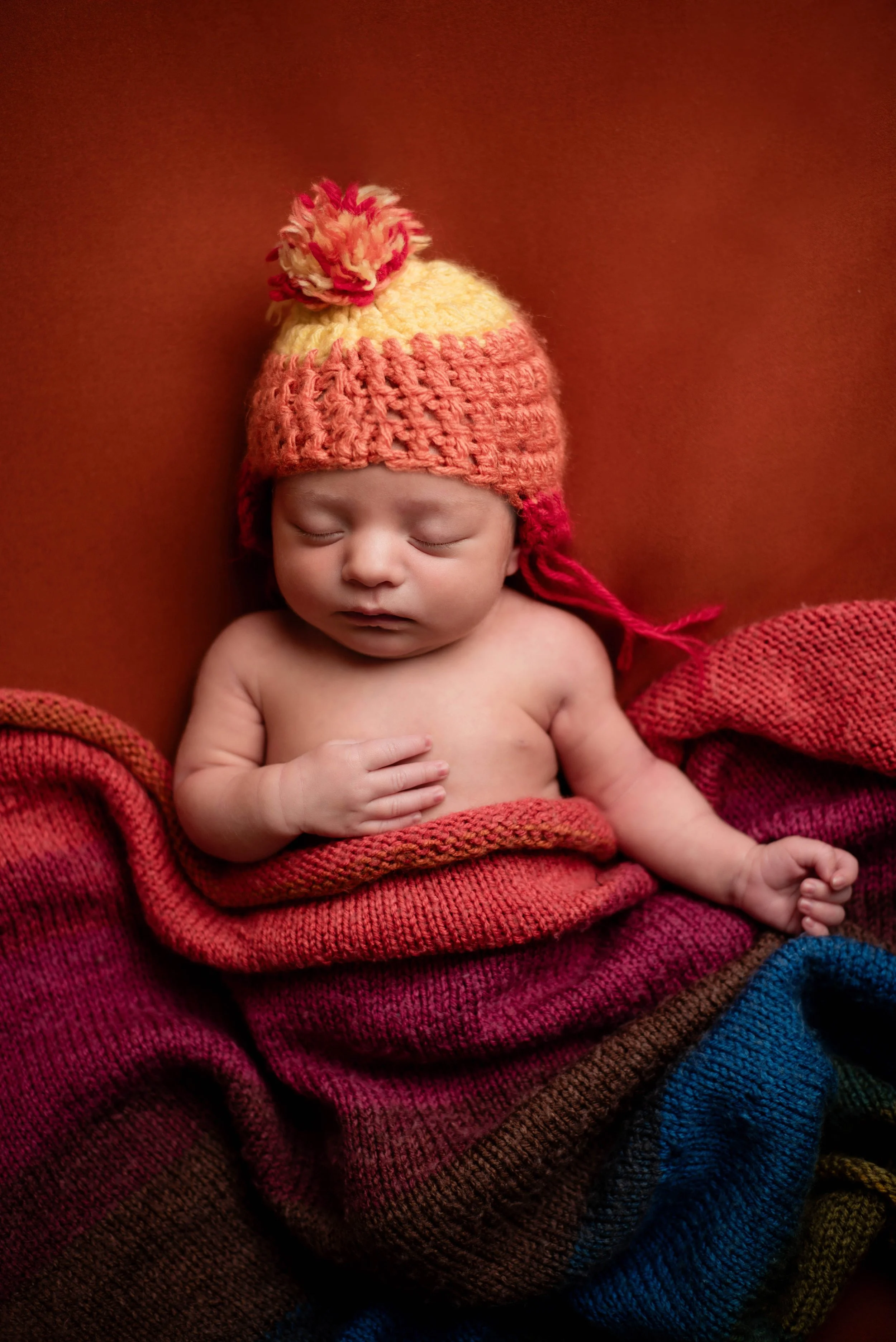 A sleeping baby wrapped in a colorful knitted blanket and wearing a rainbow-colored knitted Jayne hat with a pom-pom on top. Studio newborn portrait for Firefly nerds, Brownsville PA.
