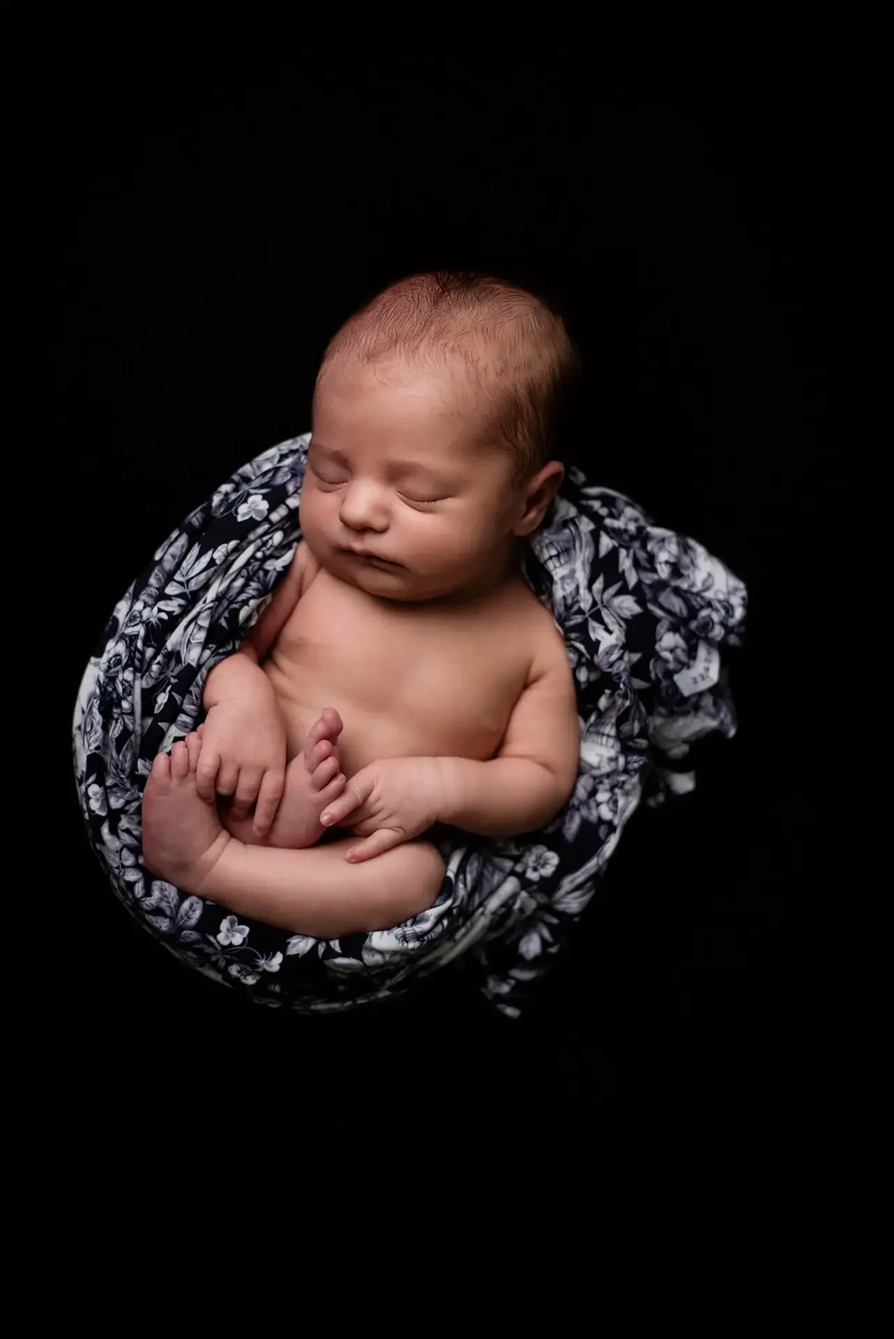 A newborn curled up on a black background, she is nested in a floral and skulls swaddle that is black and white