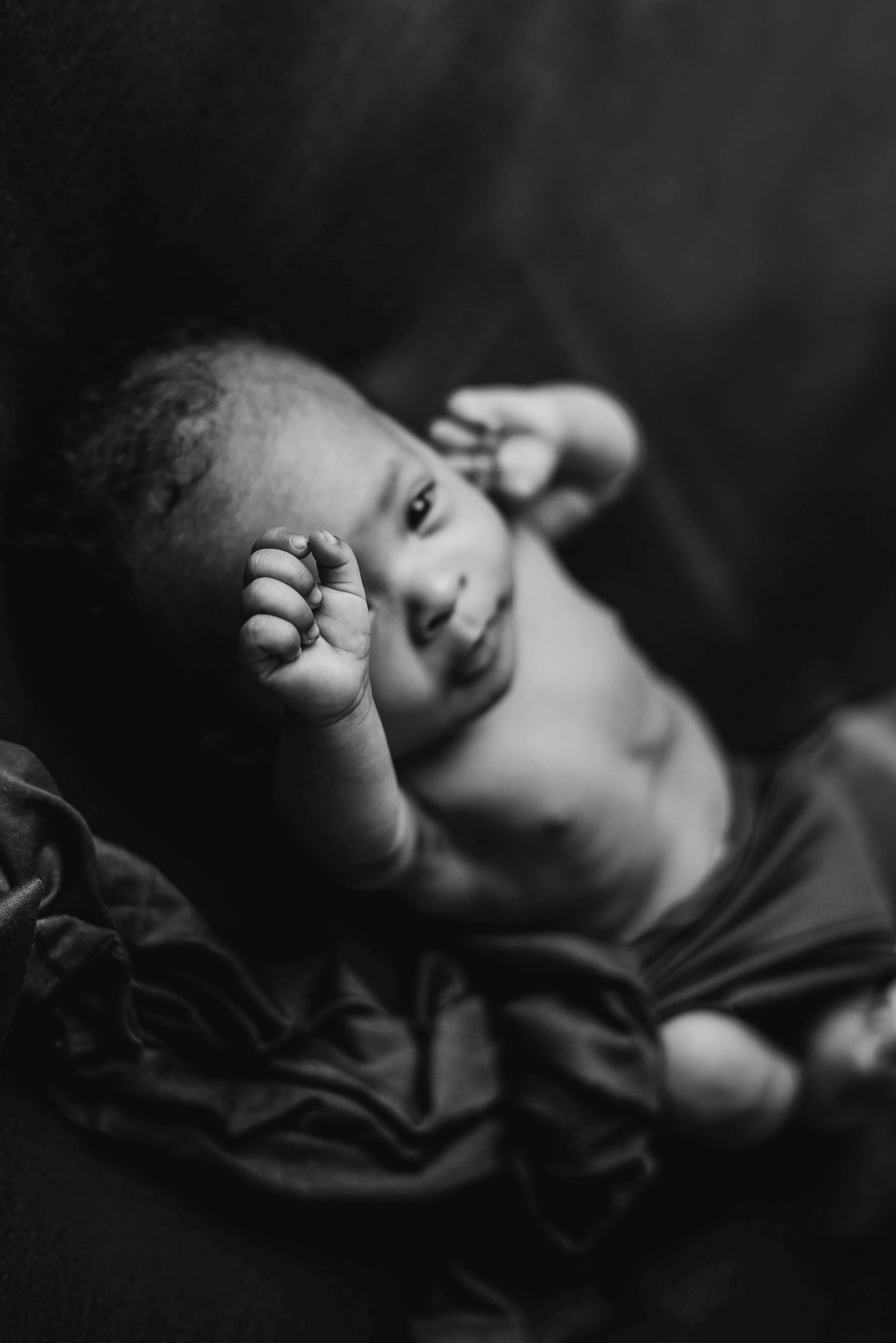 A baby lying on a dark surface, looking up at the camera with one hand near their face and the other hand raised. The photo is in black and white. Studio newborn portrait, Brownsville PA.