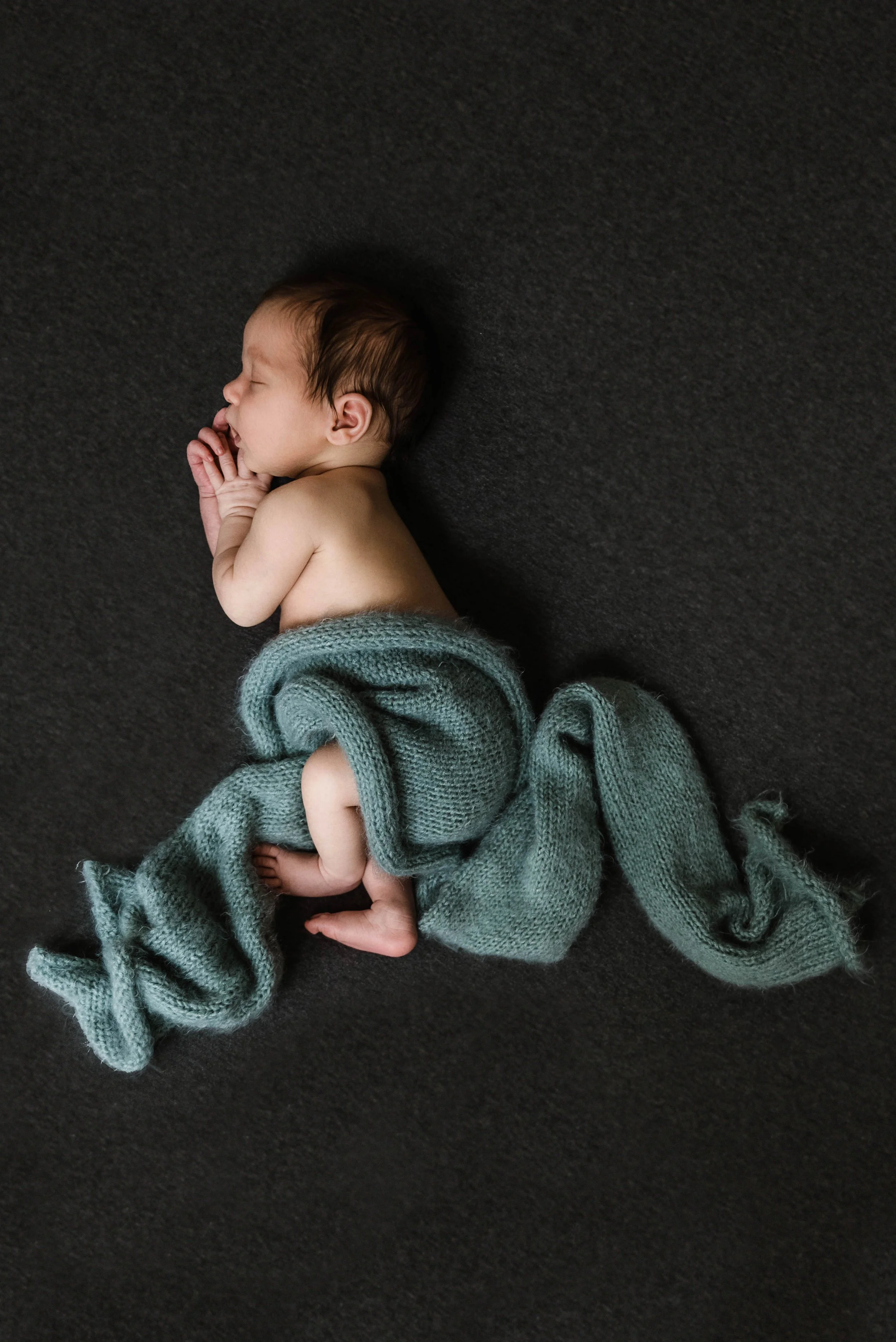 A sleeping newborn baby wrapped in a teal knit blanket on a dark surface. Studio newborn portrait, Brownsville PA.