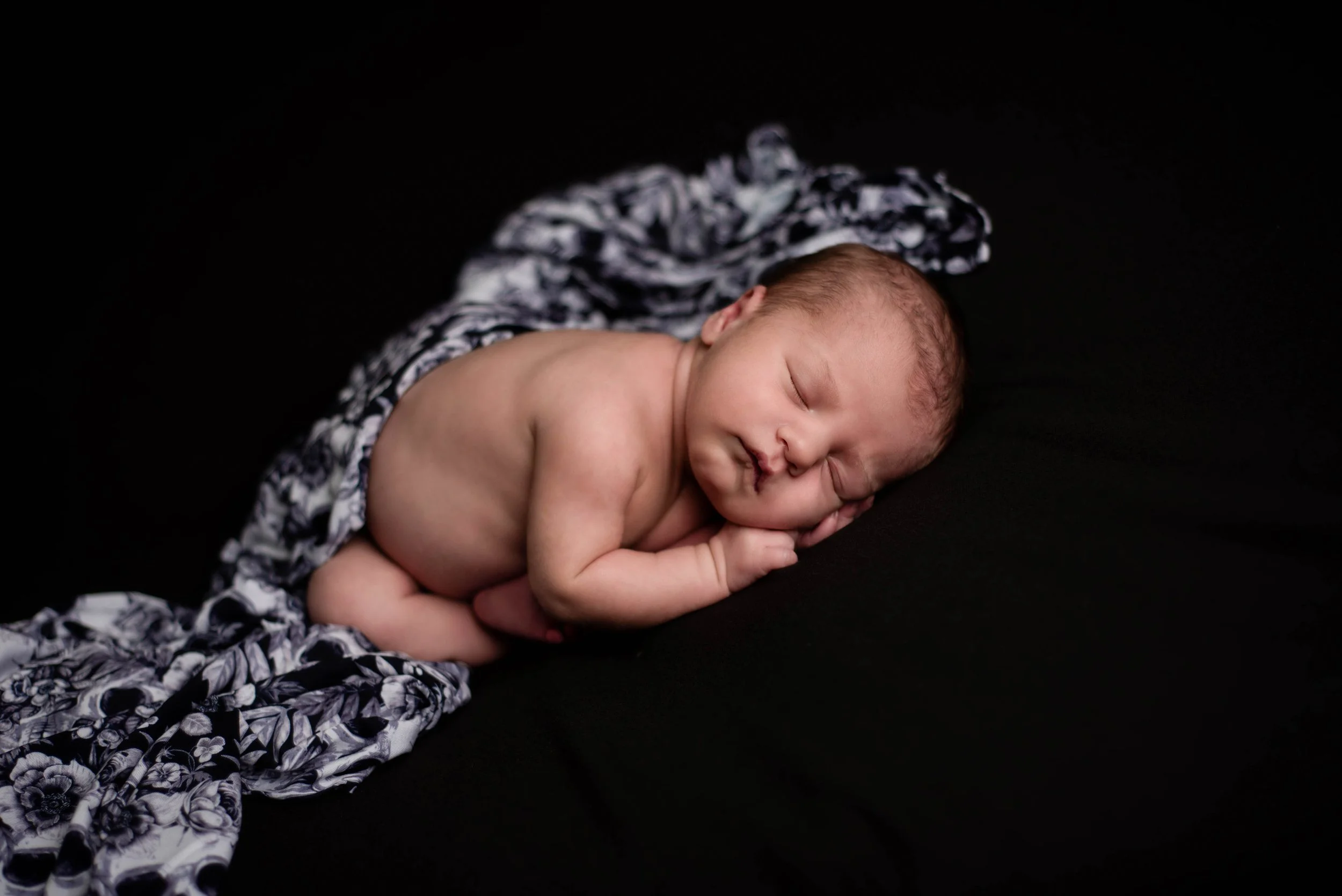 A newborn baby sleeping on a black surface, wrapped loosely in a black and white floral blanket with skulls. Studio newborn portrait, Uniontown PA.