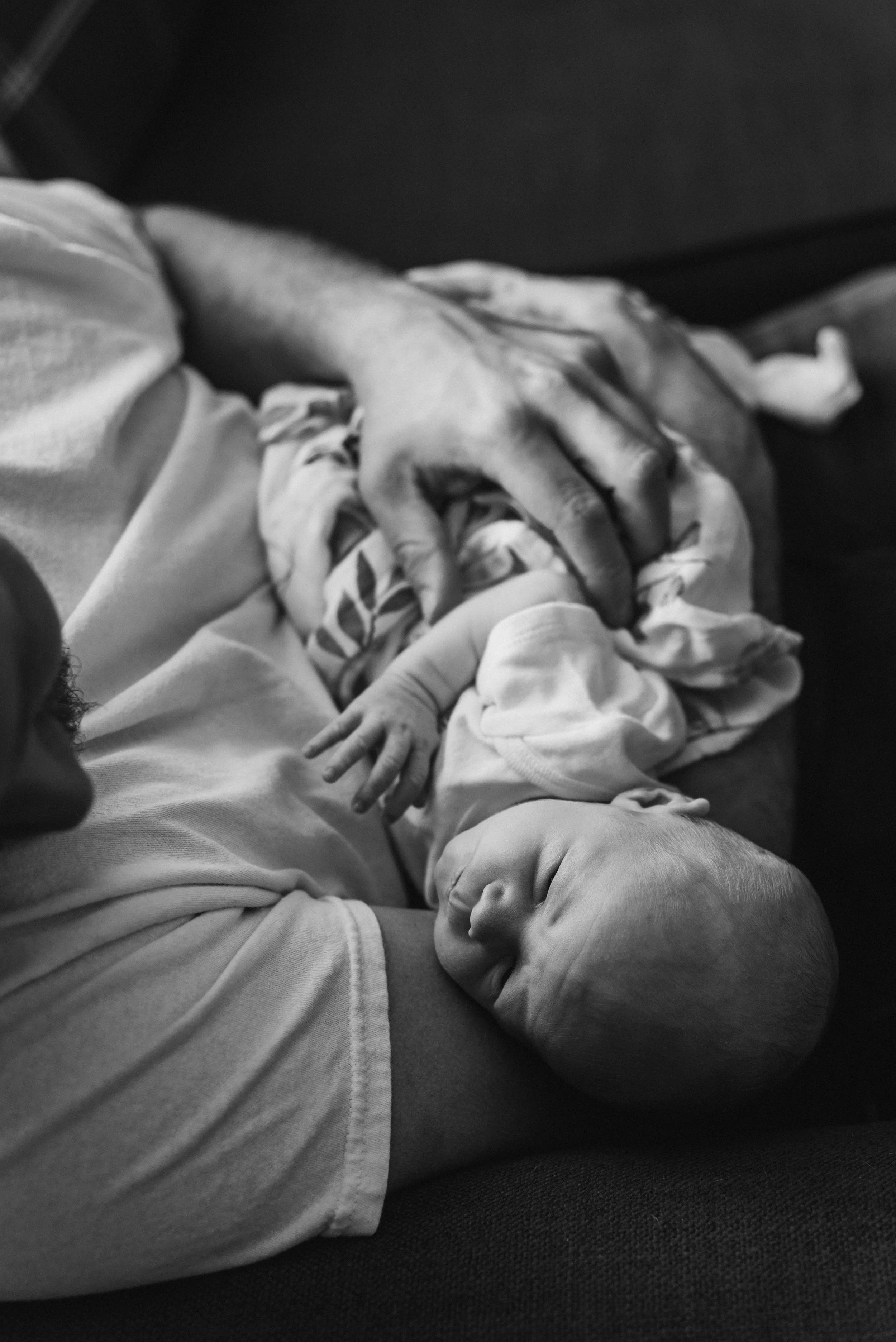 Black and white photo of a person holding a newborn baby on their chest, with one hand gently touching the baby's arm, on a dark surface. Studio newborn portrait, Brownsville PA.