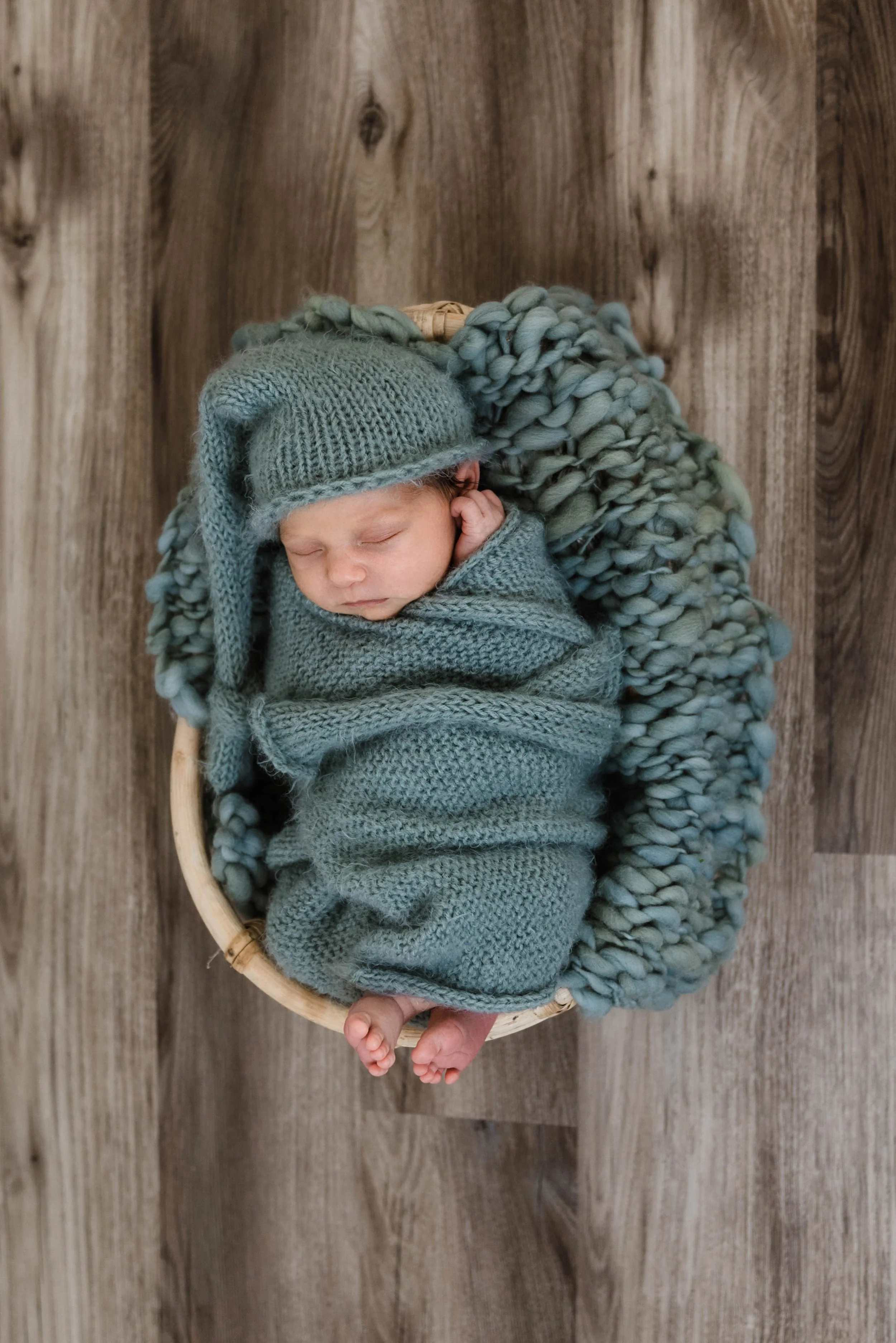 A sleeping newborn baby wrapped in a knitted blue blanket, wearing a matching knitted hat with a brim, in a round wicker basket, on a wooden floor, with a chunky blue blanket lining the basket. Studio newborn portrait, Brownsville PA.