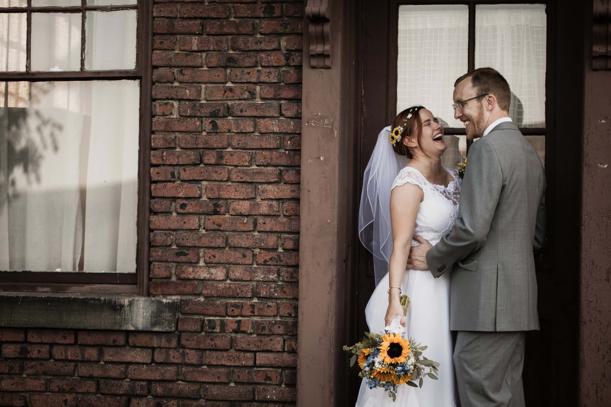 A bride and groom laugh while embracing in a doorway of a brick building at Mt. St. Macrina in Uniontown, PA