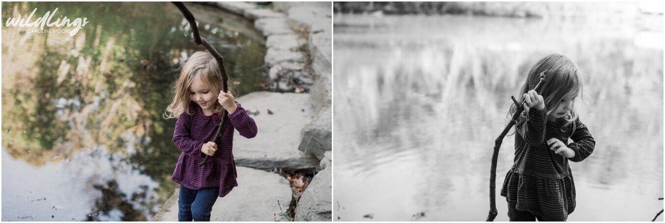 A toddler shows off her stick next to a lake surrounded by large flat stones