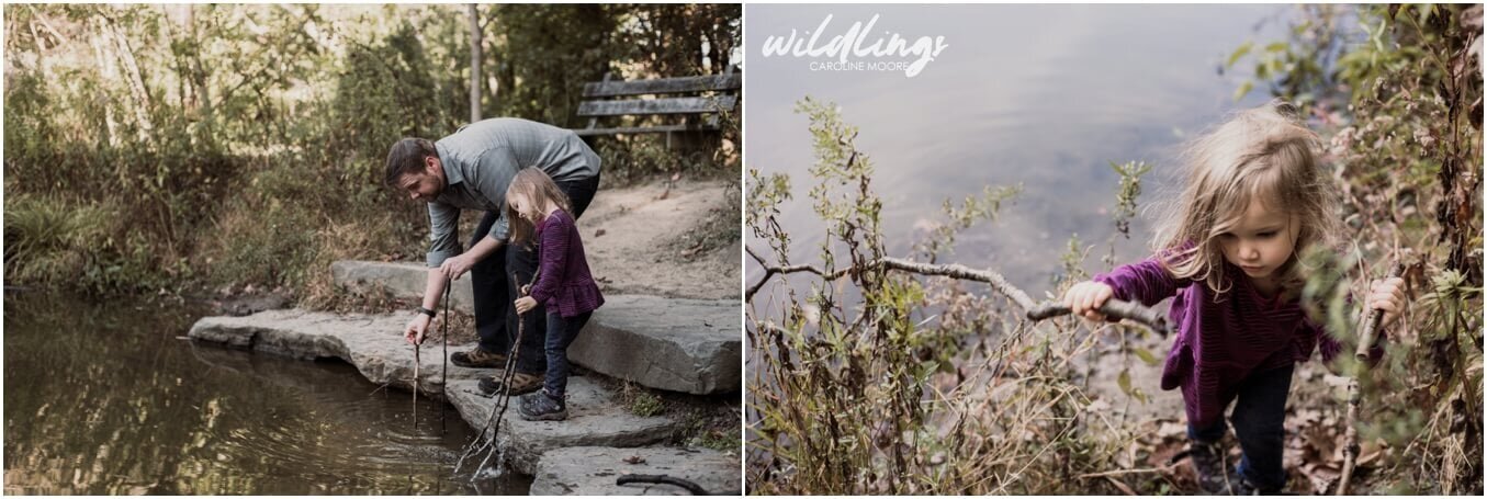 A toddler plays in the lake with sticks next to her dad, who also has sticks