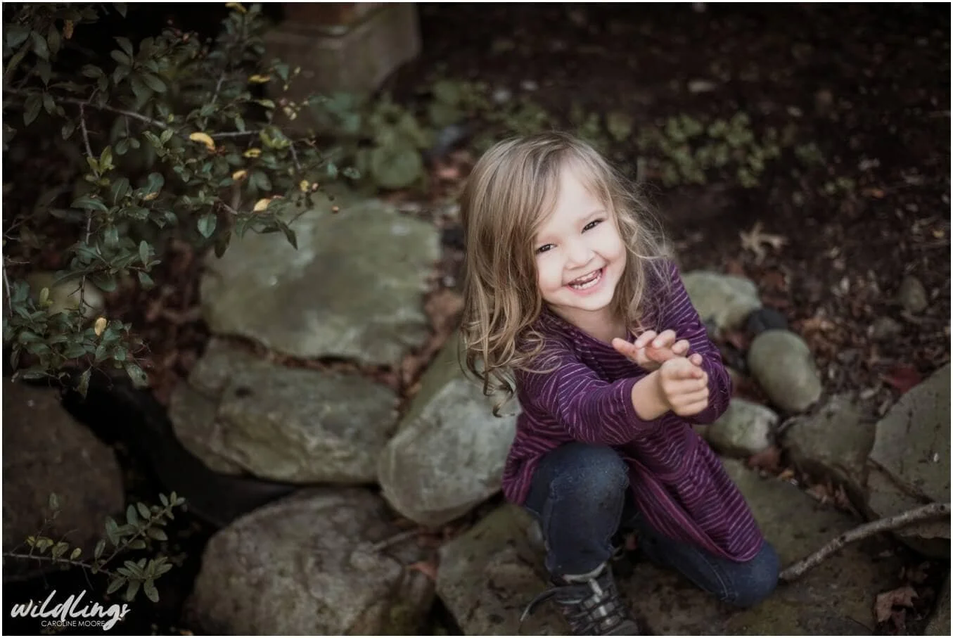 A toddler sits on large flat rocks surrounded by greenery, she is smiling at Beechwood Nature Preserve in Pittsburgh