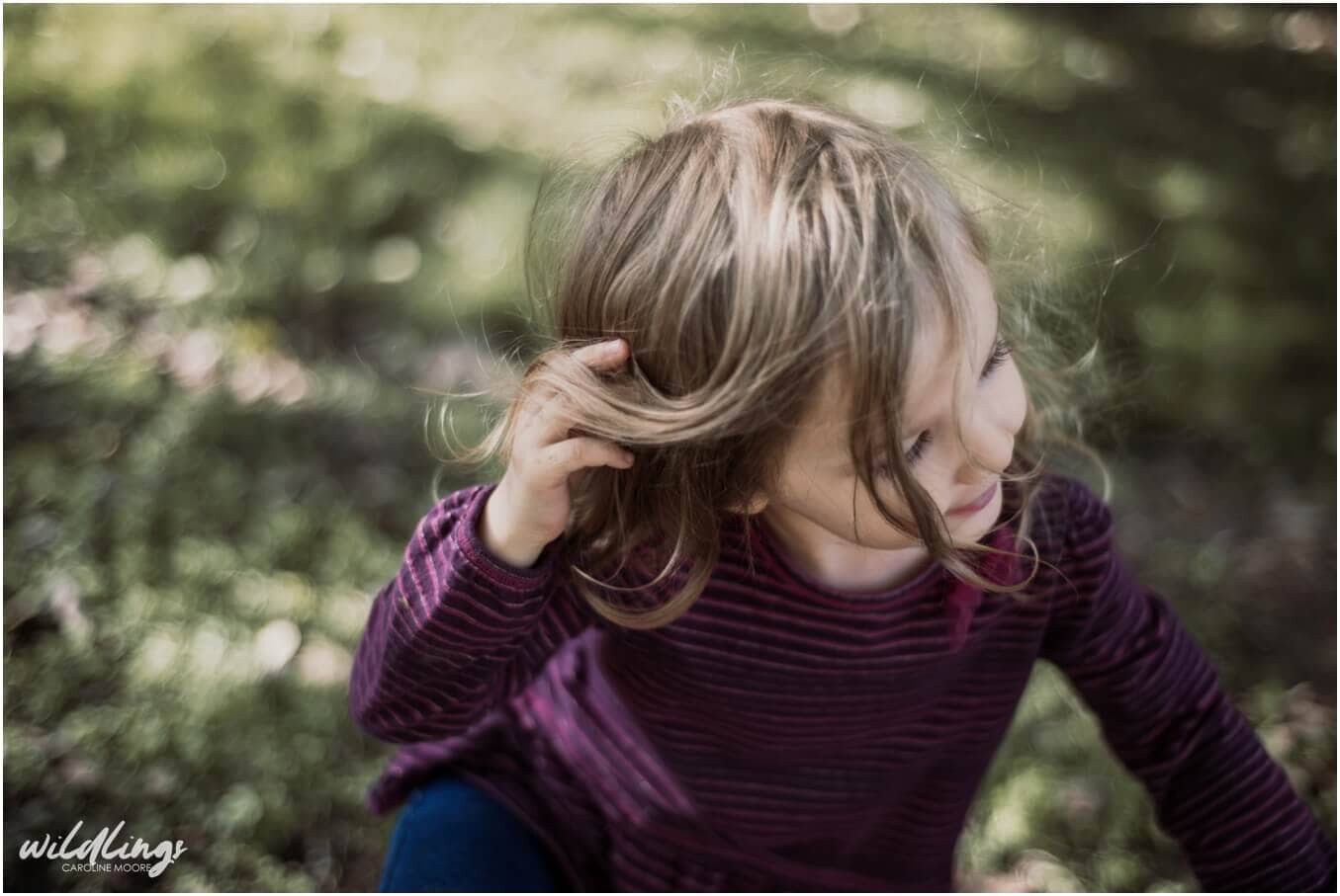 A toddler pushes her long hair out of her face, she is smiling and looking off camera