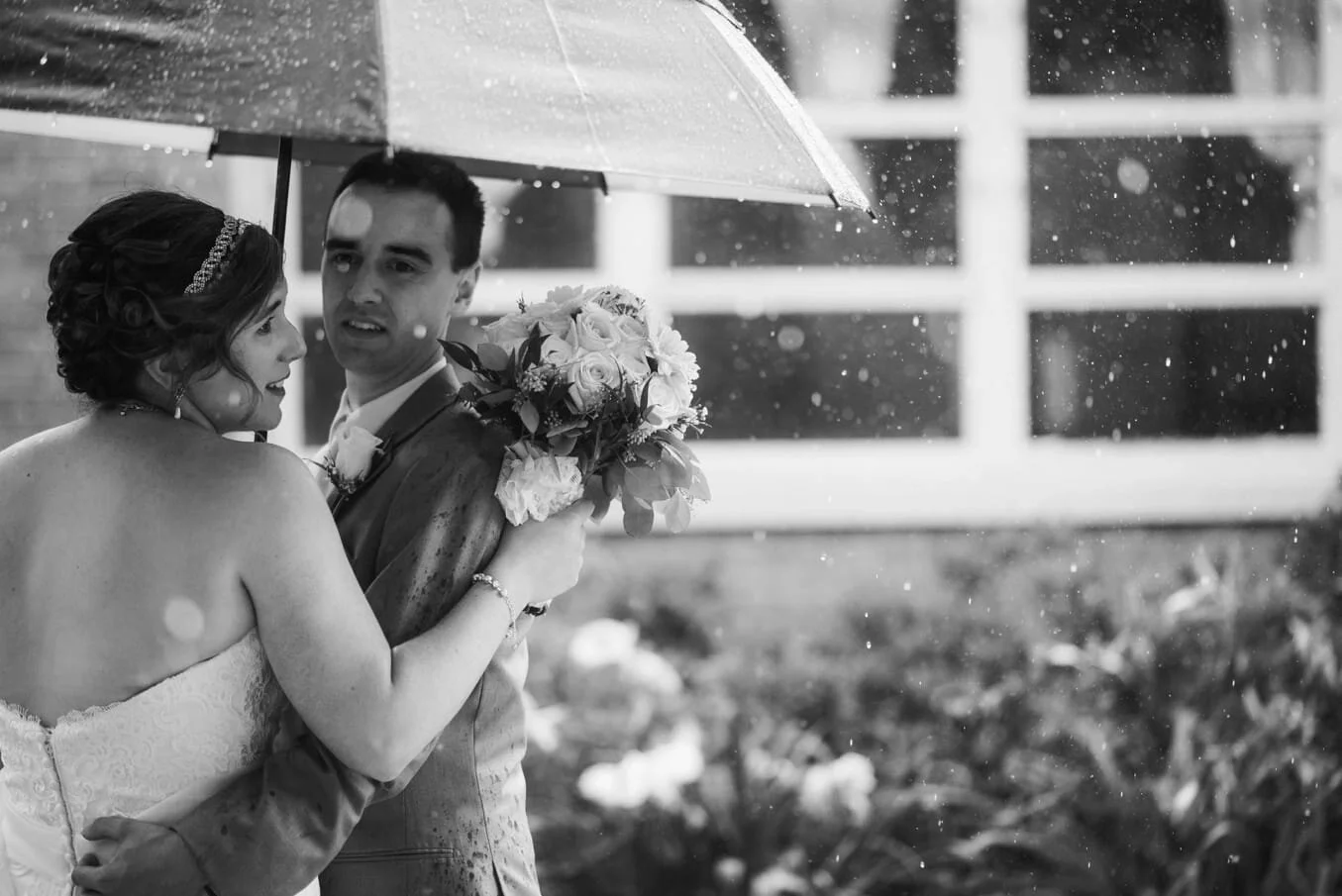 close up black and white of a bride and groom standing under a large umbrella, it is raining a lot