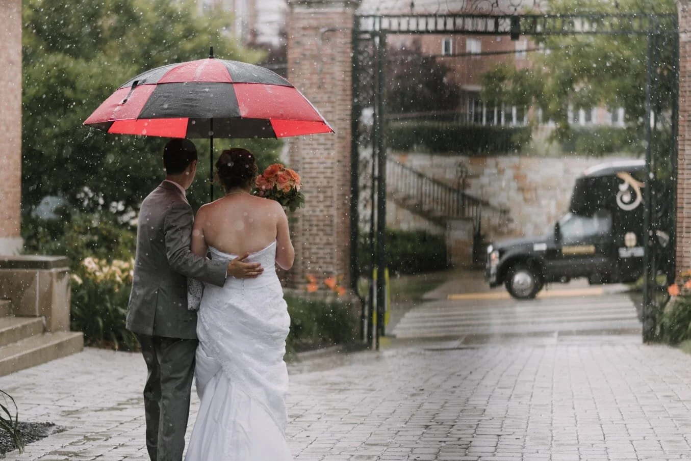 groom has his hand on bride's back as he walks her back to the party bus, he holds a large umbrella over their heads because it is raining a lot