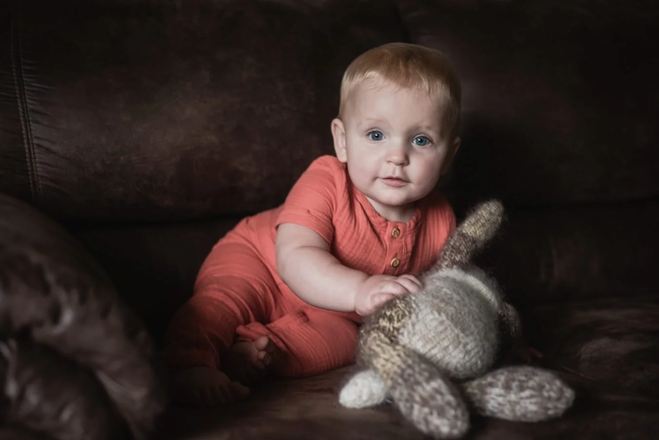 7-9 month old baby on a deep brown couch, holding a knitted bunny and looking at camera, smiling lightly