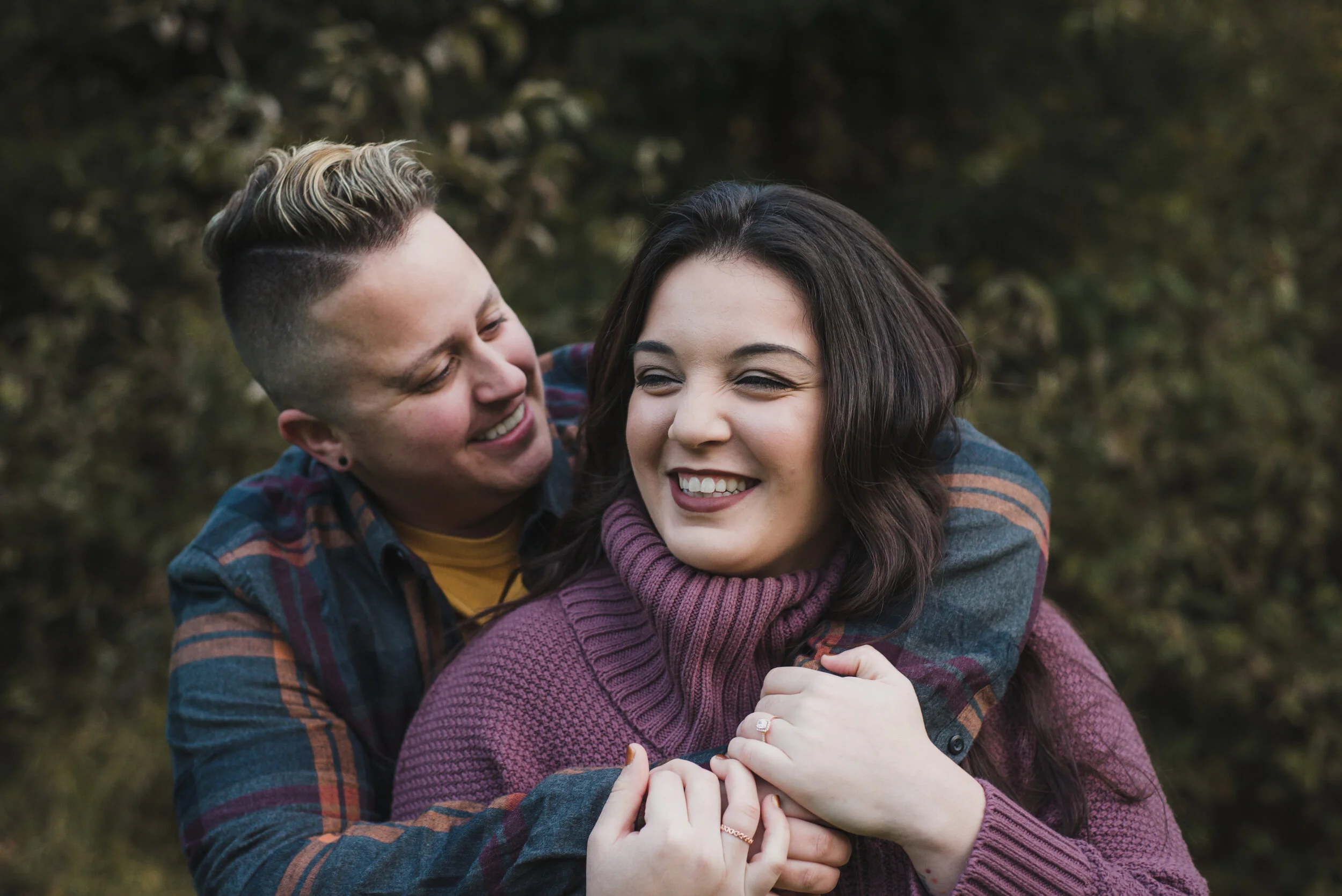 An LGBT couple during their engagement session, one woman hugs the other from behind, they are dressed for fall weather