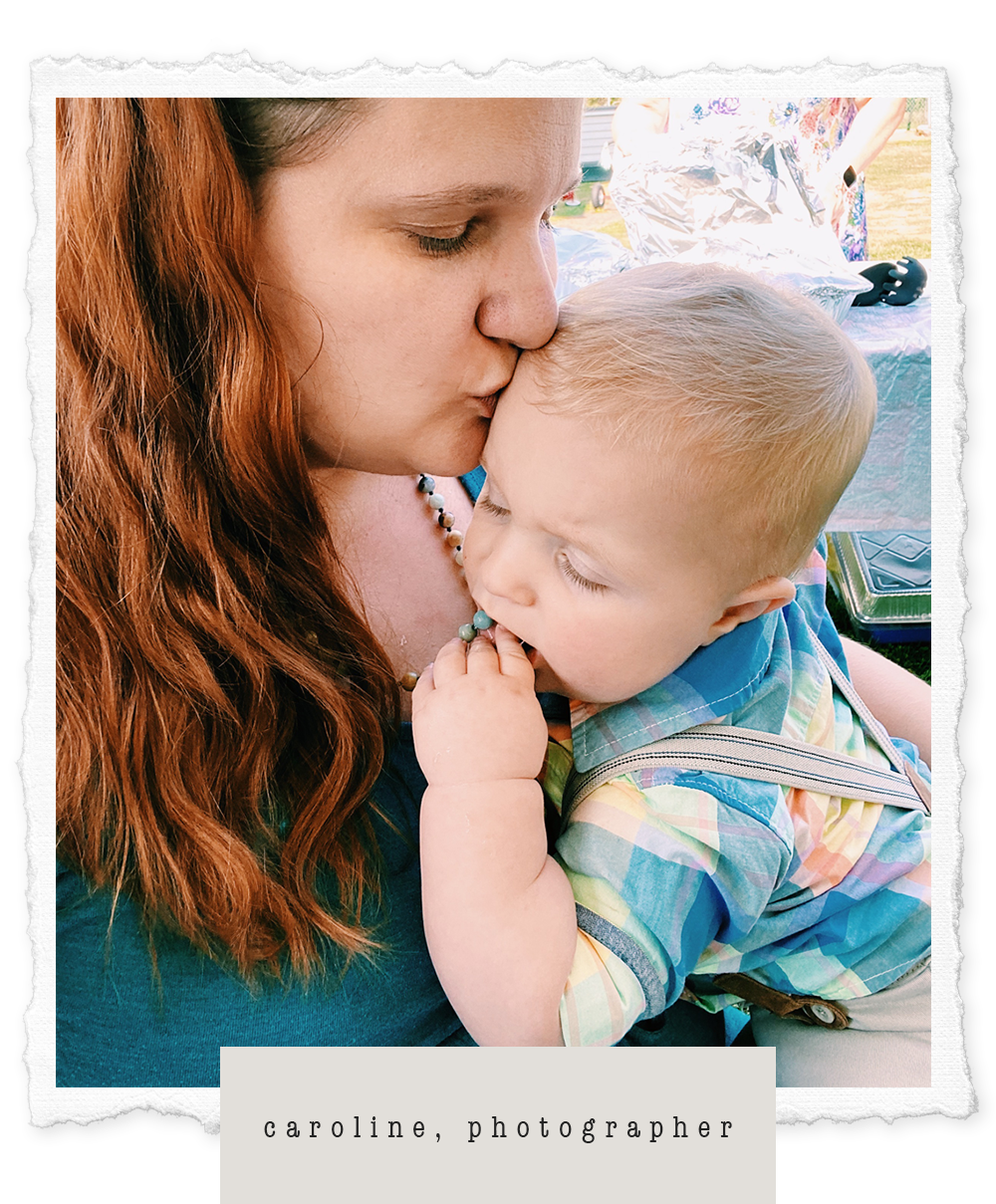 A woman with red hair kissing a young boy on the forehead, who is holding a beaded necklace and has his eyes closed, outdoors during daytime. Photographer Caroline Moore with her infant son.
