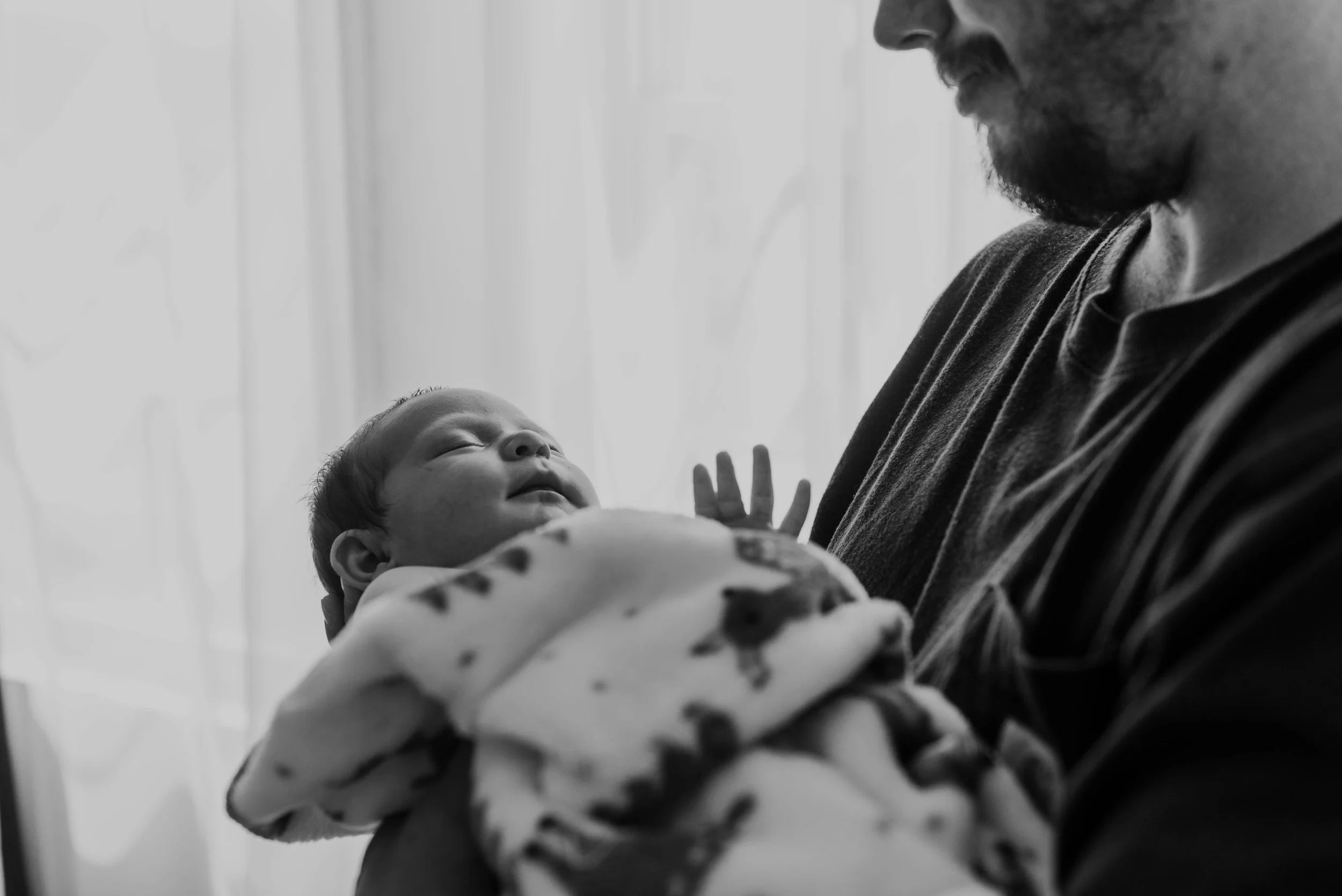 A man holding a sleeping baby in his arms, with a calm expression, near a window with sheer curtains, black and white photo. Studio newborn portrait, Brownsville PA.