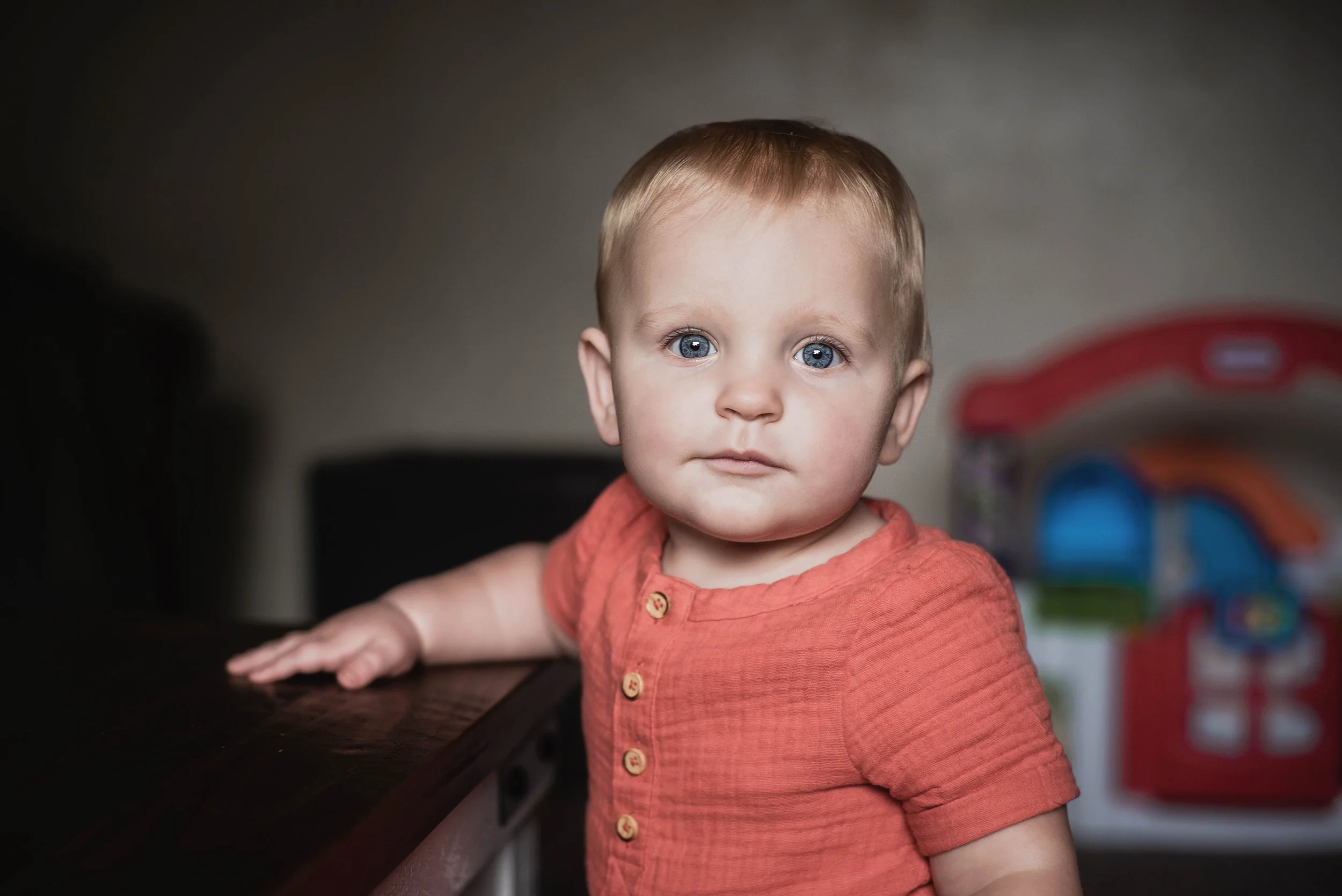A young child with blue eyes and light brown hair wearing an orange shirt, standing with one hand on a dark wooden table, looking at the camera. Milestone portraits.