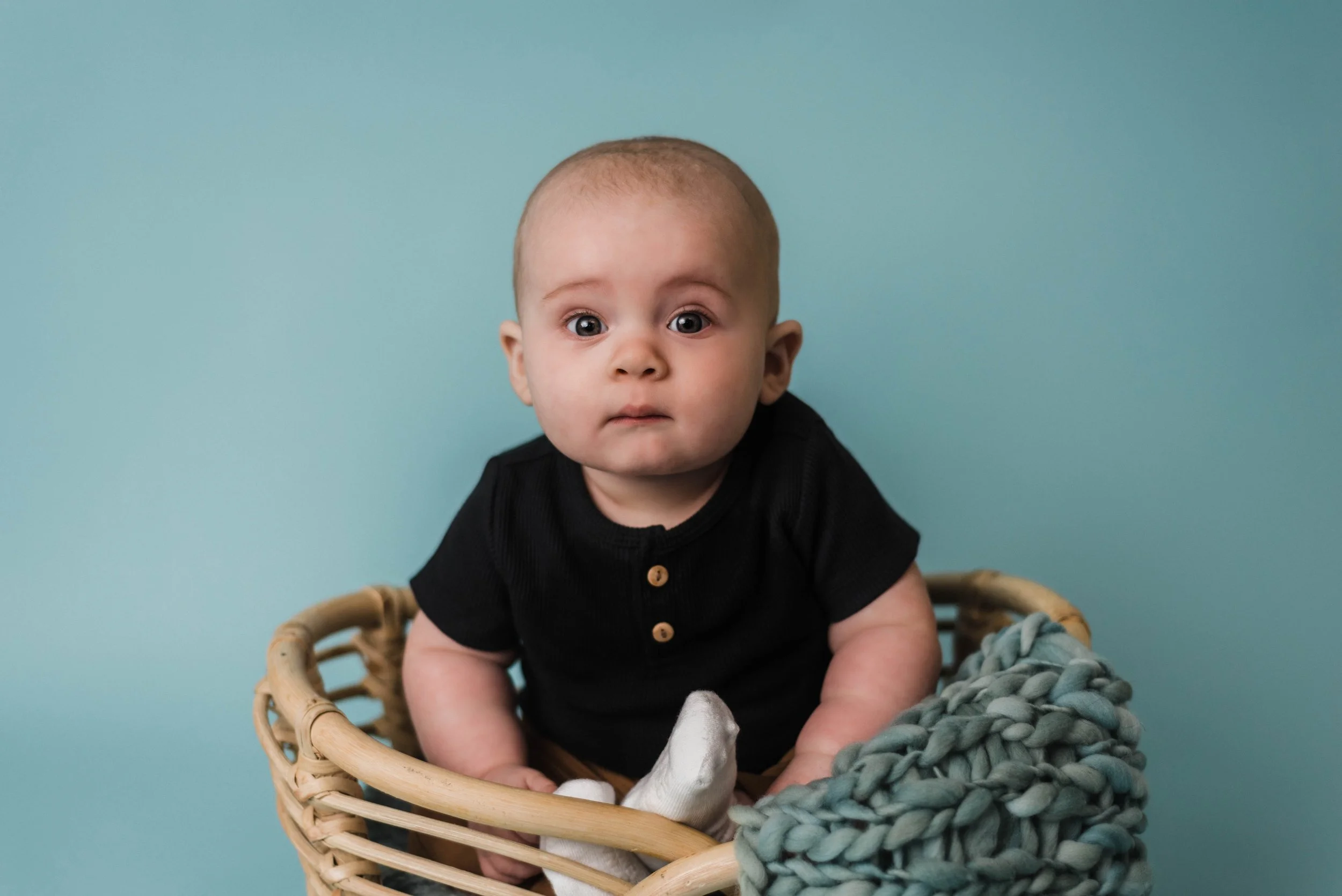 4-6 month old baby who is sitting up in a basket, he is in front of a solid blue background and is looking at camera