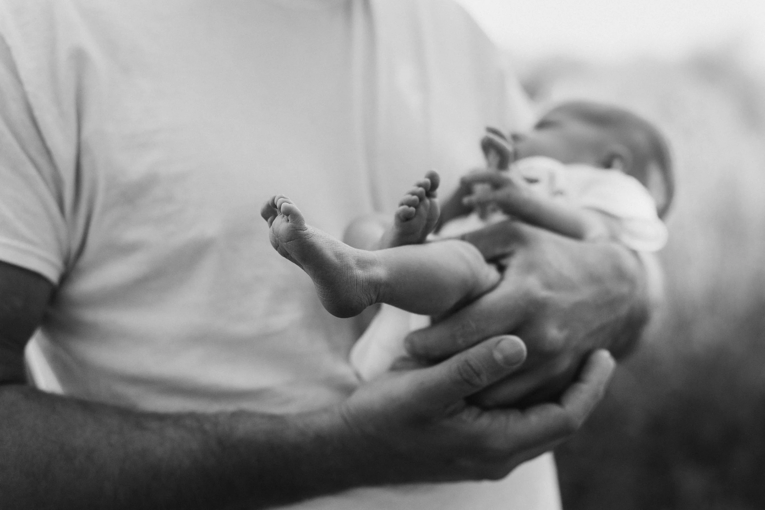 A person holding a small baby in their hands, with the baby's feet and legs prominently visible, in a black and white photograph. Studio newborn portrait, Brownsville PA.