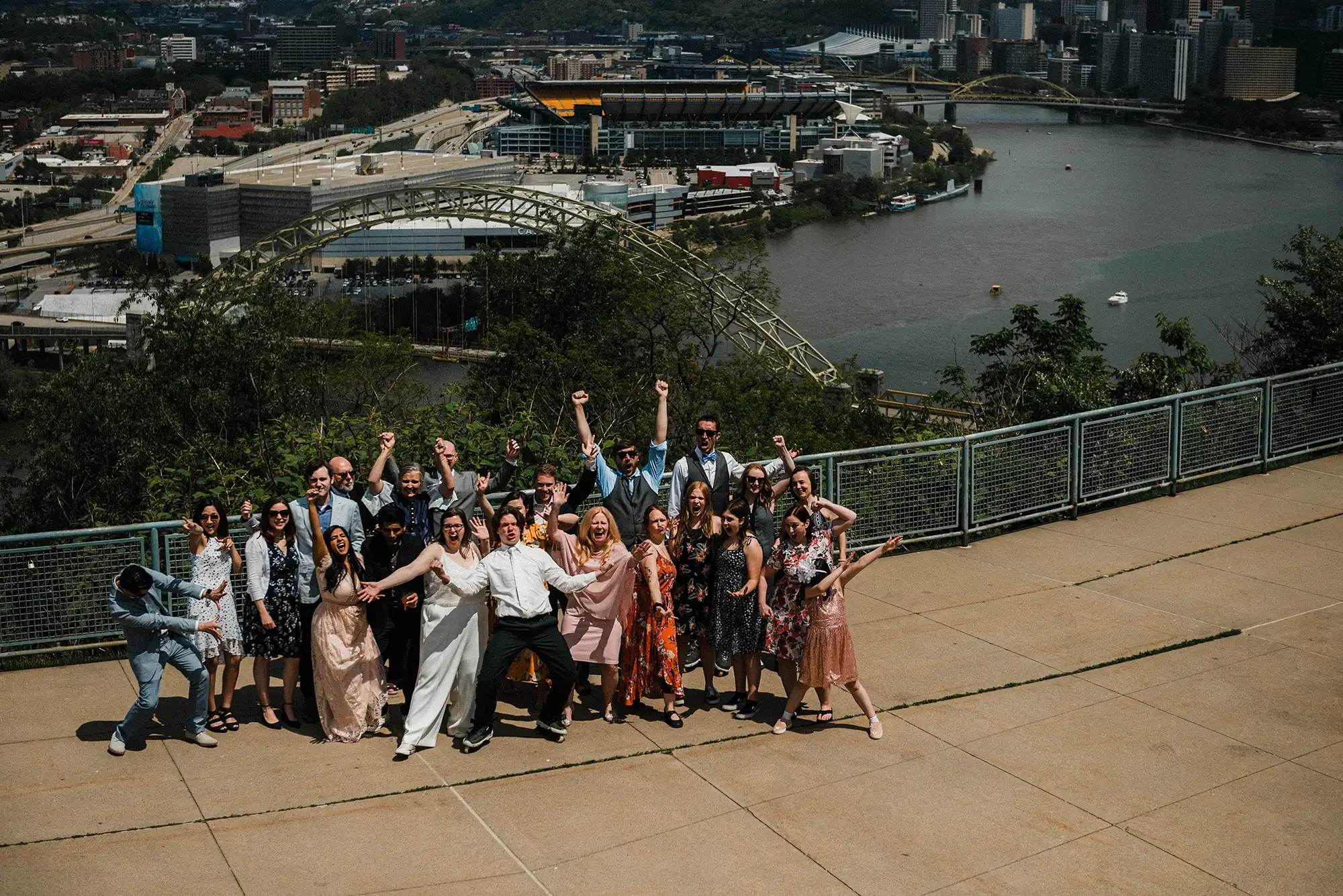 A large group of people in formal and semi-formal attire celebrating on a rooftop with a cityscape and a river in the background, some raising their arms and cheering. Photo of all the guests at a West End Overlook elopement in Pittsburgh.